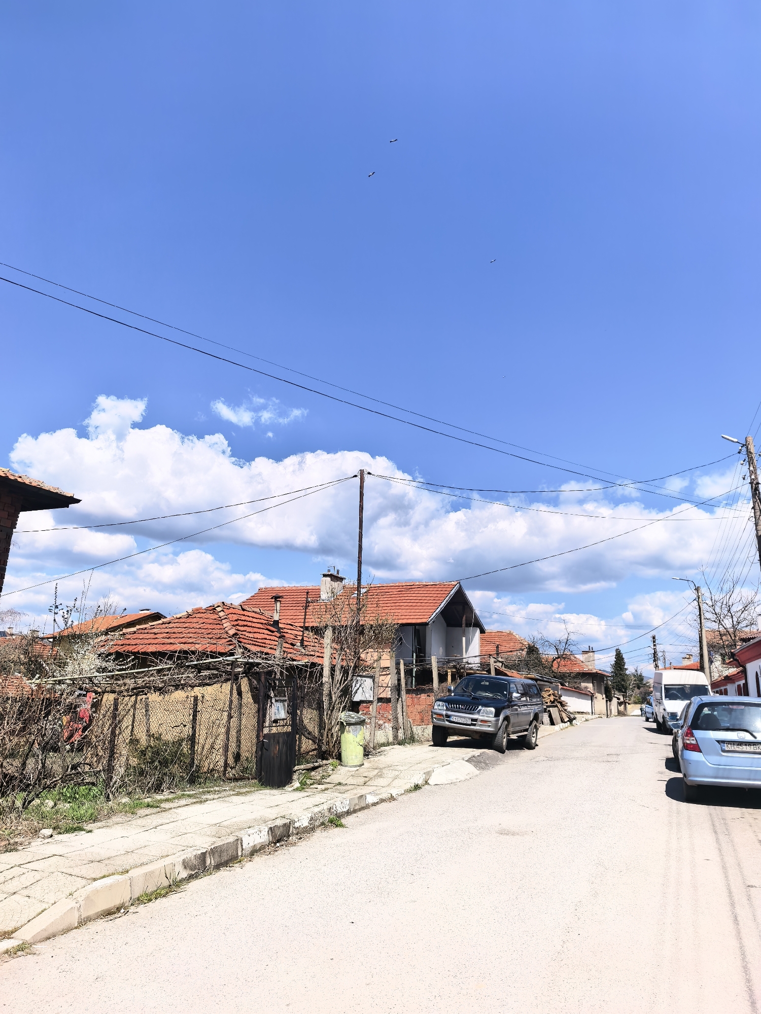 The image captures a street scene in a residential area. The sky is bright blue with scattered white clouds. Overhead, power lines stretch across the frame, and a few birds are visible against the sky.
On the left side of the street, a building with a red-tiled roof is partially obscured by a fence and climbing vines. Next to the building, a green trash can stands near a wooden gate.
A dark-colored SUV is parked on the right side of the street, partially on the sidewalk. Further down the street, several more cars and a white van are parked. The road surface is light-colored and shows signs of wear.
The overall environment suggests a small town or village setting, with modest housing and a quiet, unhurried atmosphere. The location is Unknown, Unknown.