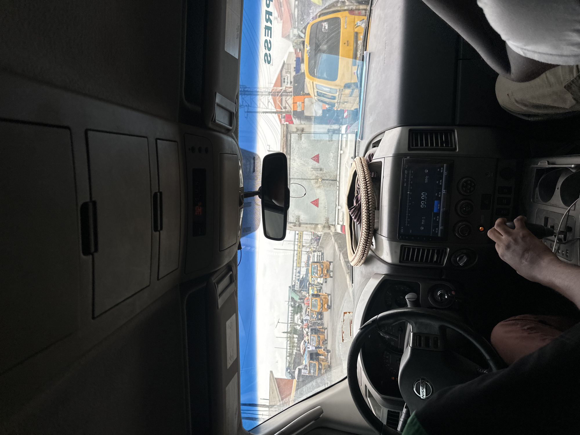 The image, correctly oriented, shows the interior of a vehicle from the passenger side, looking forward through the windshield. A person's right arm and leg are visible on the lower right, with their hand on the gear shifter. Part of another person's body is in the upper right corner. The vehicle's interior features a dark dashboard, a Nissan-branded steering wheel, a rearview mirror, and an infotainment system.
Outside, through the windshield, numerous yellow three-wheeled auto-rickshaws (commonly known as kekes) are queuing on what appears to be a busy street or road. A larger yellow vehicle, possibly a bus or truck, is partially seen on the left side of the windshield view. Buildings, scaffolding, and power lines are visible in the background.
The scene is brightly lit by daylight, suggesting daytime. The concentration of yellow auto-rickshaws is a strong visual cue for a West African urban setting, consistent with Isheri, Nigeria. The vehicle appears to be stationary or moving slowly in traffic.
Visible text includes the "NISSAN" logo on the steering wheel. The infotainment system displays "99.90 MHz", "FM 1", and "PM 1". Reflected on the top-center of the windshield, the word "PRESS" is visible in reverse ("22SSƎЯP"). Additionally, two red triangular hazard symbols are visible on white backgrounds outside the vehicle.
