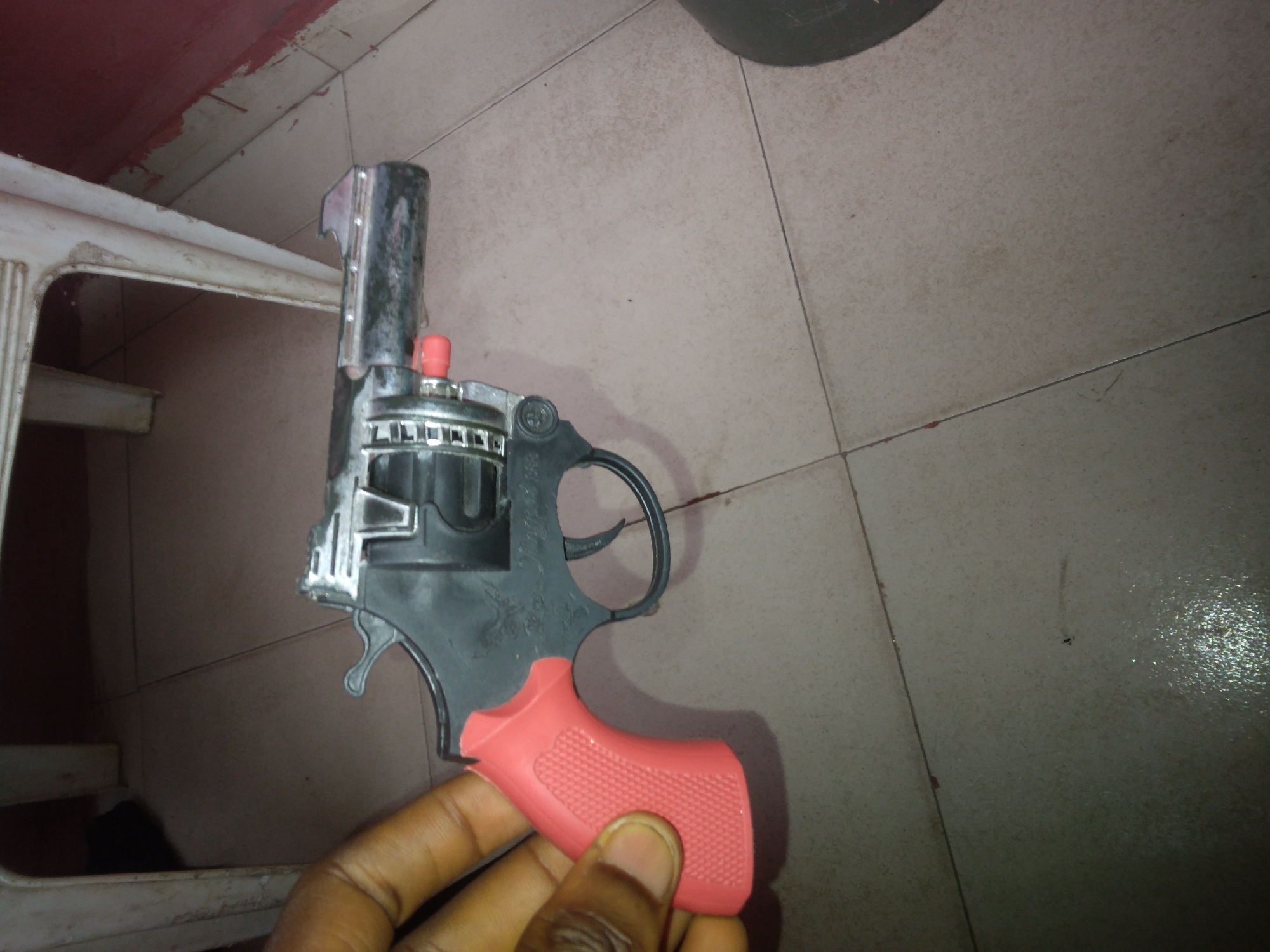 A black and silver toy revolver with a red grip is held in a person's hand. The background shows beige tiled flooring and part of a white plastic chair. A dark object is visible in the upper right corner. The location is inferred to be Onitsha, Nigeria based on the user prompt.