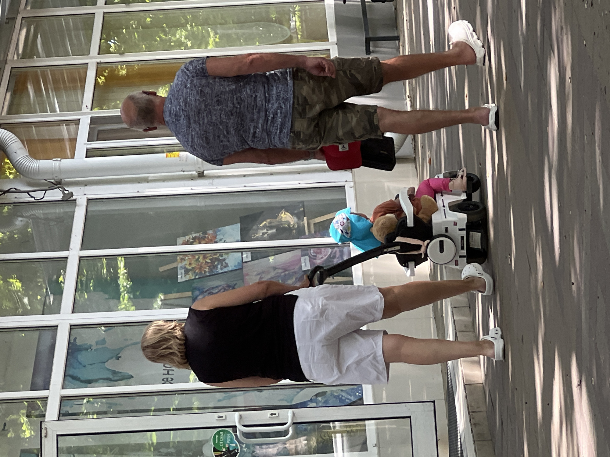 The image depicts a street scene in Krasnodar, Russia, on a sunny day. Two adults, a woman and a man, are walking away from the camera. The woman, in the foreground and to the left, wears a black sleeveless top and white shorts. She has blonde hair and is pushing a white toy car-shaped stroller. The man, to the right, wears a grey patterned t-shirt and camouflage shorts. He is carrying a red and black bag. Inside the stroller, a child wearing a blue hat and pink pants is seated, along with a brown stuffed toy. They are walking on a paved walkway in front of a building with large glass windows, through which paintings and greenery are visible. The environment suggests an outdoor area, possibly near an art exhibition or a public space.