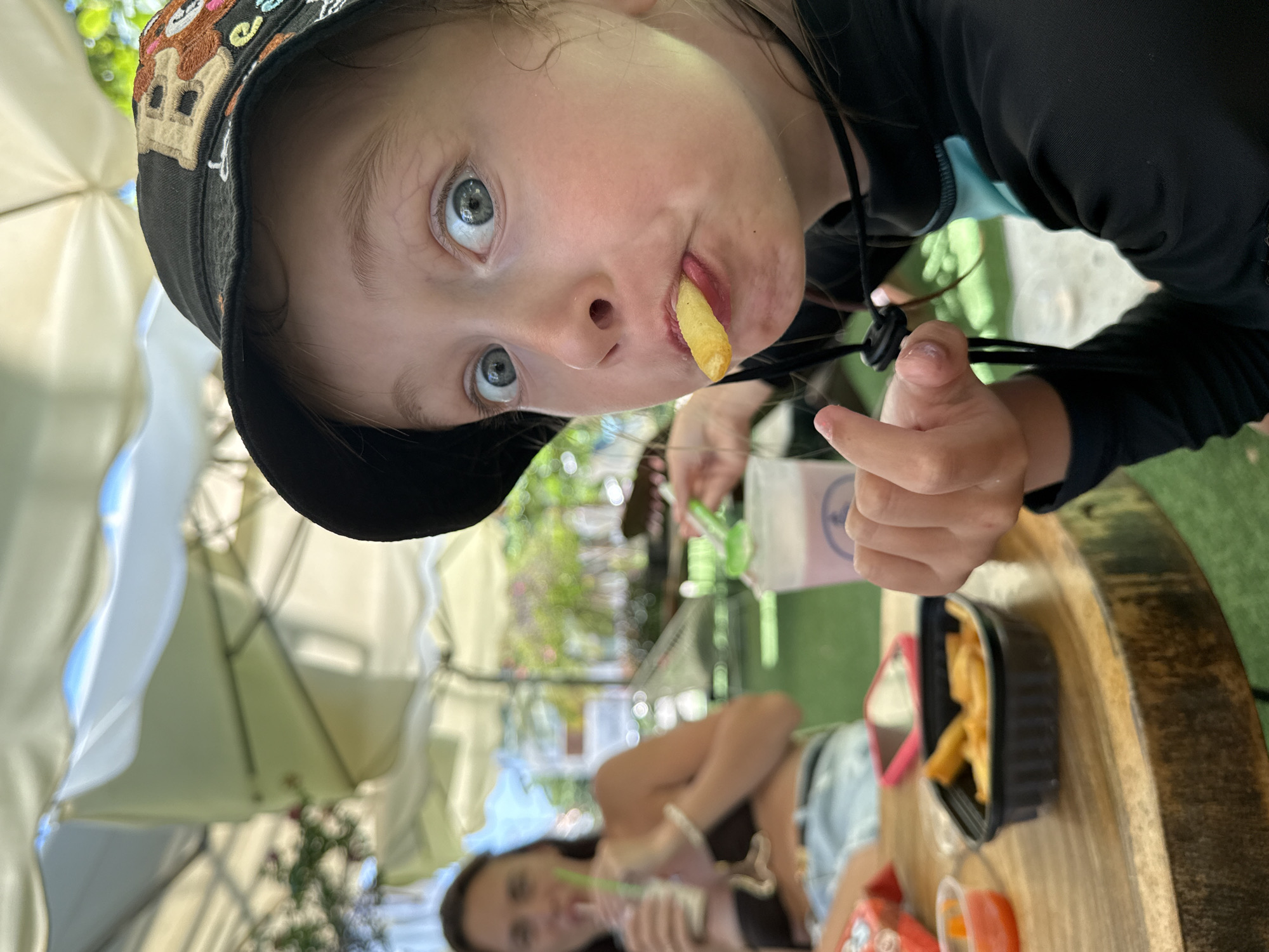 A close-up shot shows a young child wearing a black hat with a cartoon bear design. The child is holding a french fry in their mouth and looking at the camera with wide, blue eyes. The child's face is illuminated, suggesting it's daytime. The background is blurred but shows an outdoor setting with people, umbrellas, and greenery.
To the right, a woman is seated, and another person, possibly an adult, is visible in the background, with their arm resting on a table. There are containers of food, including french fries and dipping sauce, on the table. The setting appears to be a casual dining or outdoor cafe, possibly in Nha Trang, Vietnam, given the location context. The overall mood seems relaxed and informal, with the child's playful expression being a focal point. The lighting and shadows suggest a bright, sunny day.