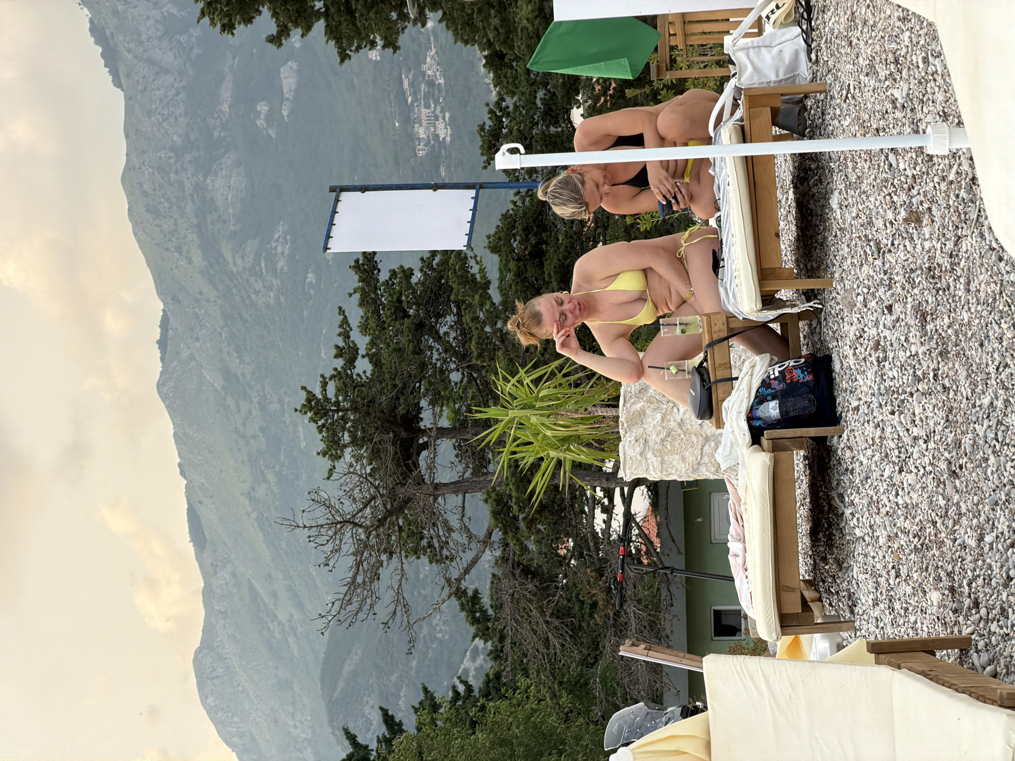 Two women are seated on lounge chairs on a pebble beach. The woman on the left is wearing a yellow bikini and leaning on her hand. The woman on the right is wearing a black bikini and looking at her phone. In the background, there are trees and mountains. A white sign on a blue post is visible. A green flag is hanging on a wooden structure to the right. The scene appears to be in Budva, Montenegro.