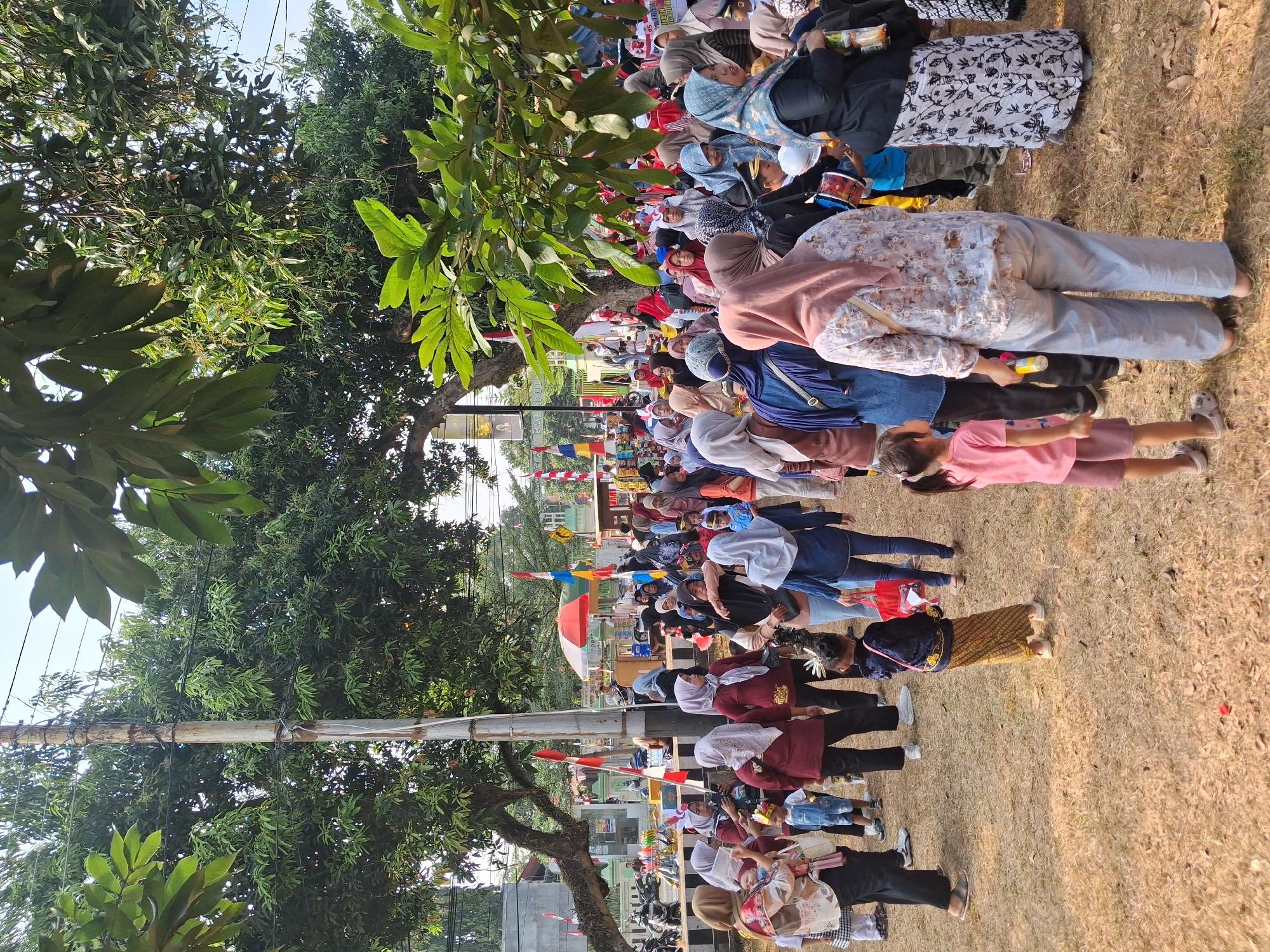 In Balapulang, Indonesia, a crowd of people, predominantly women and children, are gathered outdoors on a dry, grassy area under a canopy of trees. Many of the individuals are wearing headscarves or hijabs. Some are dressed in traditional attire, including sarongs and head coverings adorned with decorative elements. In the background, several red and white flags are visible, along with what appears to be a small market stall or food vendor area with a red umbrella. The overall atmosphere suggests a local event or celebration.