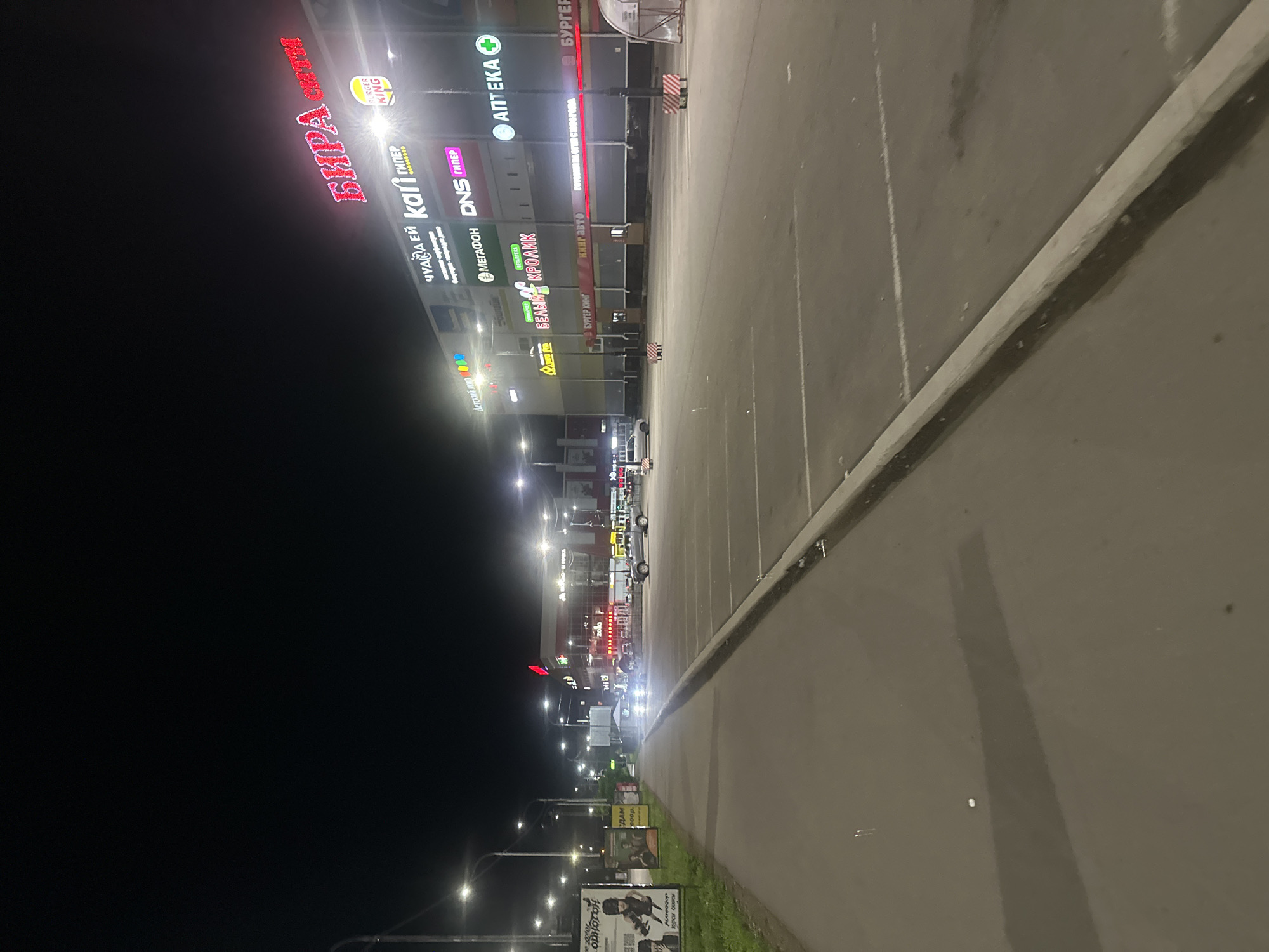 This is a nighttime, exterior, vertical, eye-level shot of a shopping center facade in Birobidzhan, Russia. The shopping center features multiple bright, colorful signs advertising various businesses, including "БИРА" (Bira), "BURGER KING," "АПТЕКА" (Apteka), "DNS," "МЕГАФОН" (Megafon), and "Детский мир" (Detskiy mir). The scene is a dimly lit parking lot in front of the shopping center. The parking lot is mostly empty, with a few cars visible. The lighting suggests it is nighttime, and the presence of streetlights and illuminated signs confirms this. The weather appears to be clear. The overall mood is commercial and vibrant due to the illuminated signs, but also quiet and somewhat desolate due to the empty parking lot and nighttime setting. The image is slightly blurry, possibly due to low light conditions or camera movement. Several signs are visible and legible, providing information about the businesses located within the shopping center.