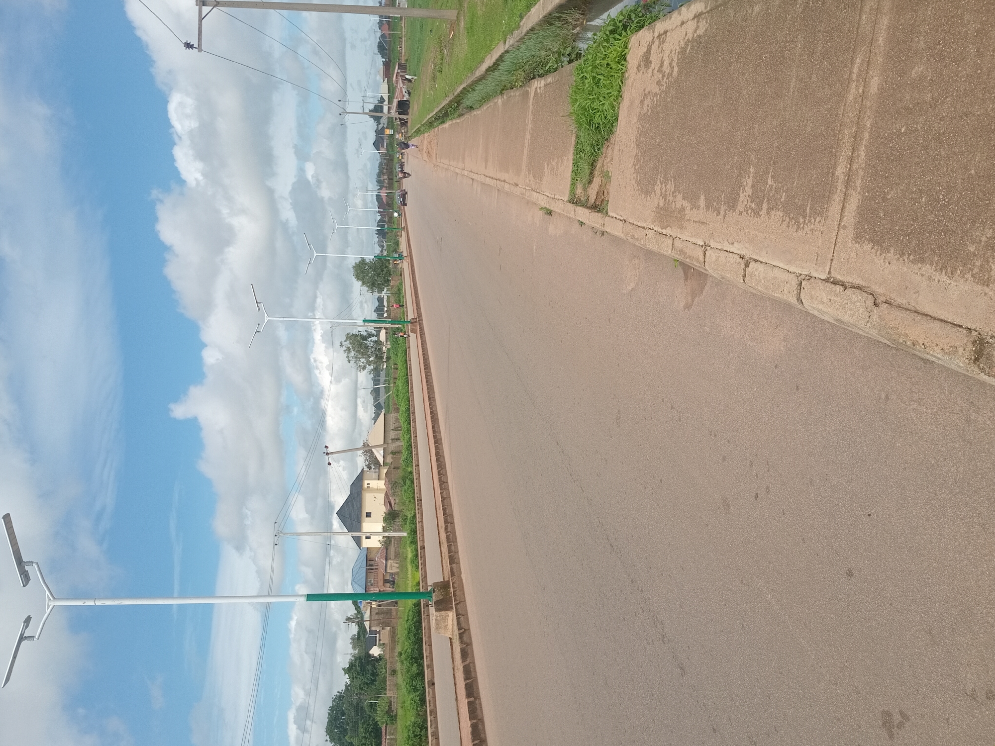 The image captures a roadside scene in Bukuru, Nigeria. A paved road runs diagonally across the frame, with a concrete sidewalk on the right. Several solar-powered streetlights line the left side of the road, some with green bases. In the background, houses and buildings are visible, interspersed with trees and vegetation. A few people and vehicles, including motorcycles and a car, are present on the road and sidewalk. The sky is partly cloudy, with white clouds against a blue backdrop. A drainage ditch runs alongside the sidewalk.