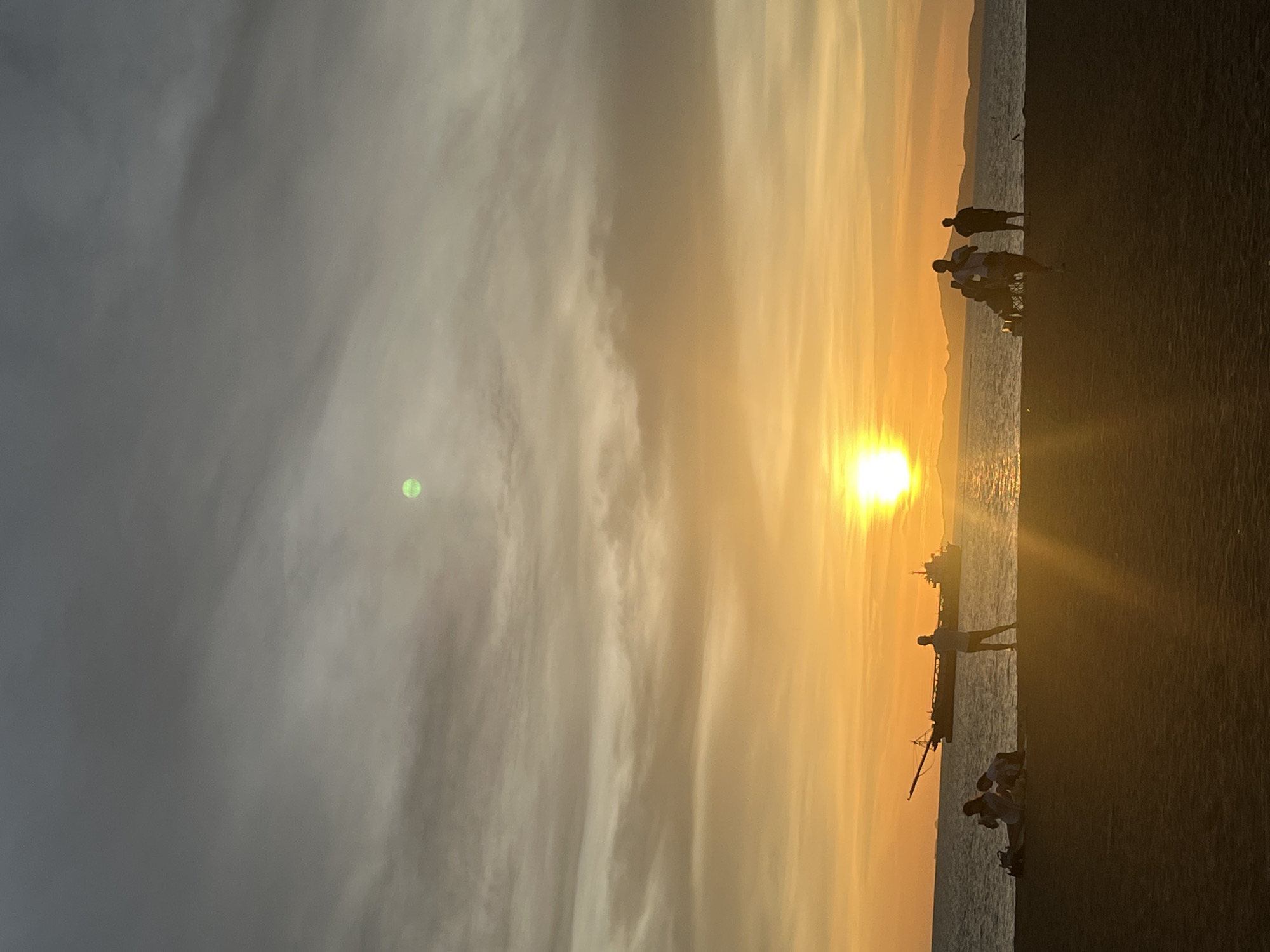 The image depicts a sunset scene at a beach in Pattaya City, Thailand. The sun is positioned near the horizon, casting a warm, golden glow on the sea and sky. Several people are visible in silhouette on the beach. A ship is visible on the sea near the sun. The sky is filled with clouds. The image is taken from a low angle, with the beach occupying the bottom portion of the frame.