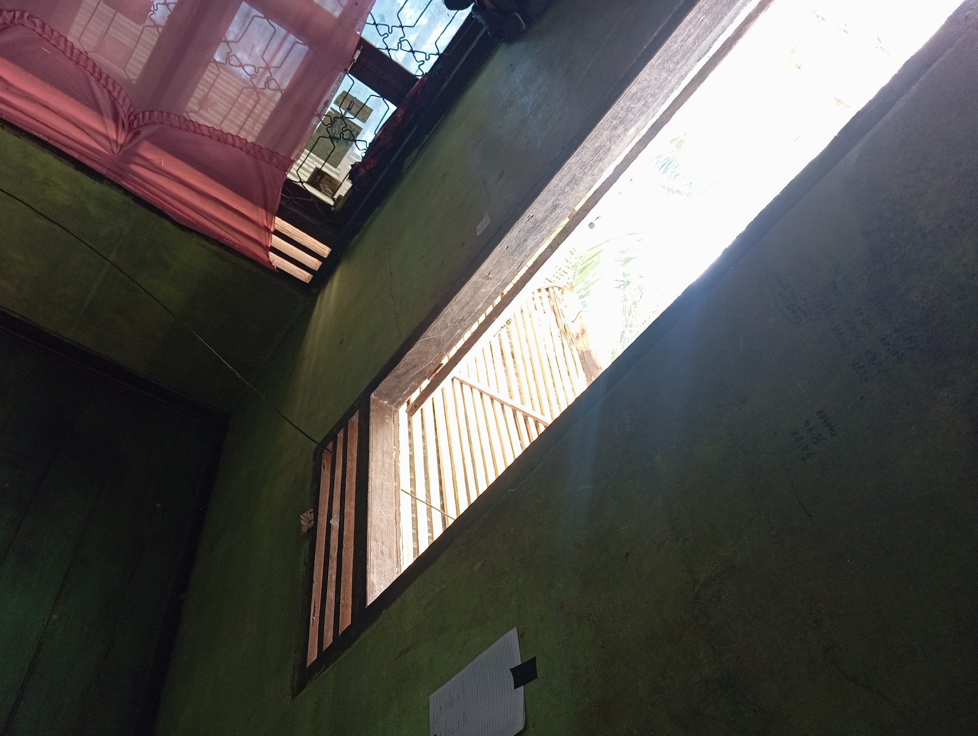The image shows a close-up, high-angle view of an interior space with a green-painted wall and wooden ceiling. A narrow, horizontal window opening, covered by wooden slats, runs across the center of the frame, allowing bright sunlight to stream in. The light illuminates the slatted window and casts shadows. To the right of this opening, a second, smaller window is partially covered by a sheer, pink curtain with a ruffled edge. The curtain is drawn back, revealing a glimpse of the window behind it. Some faint writing is visible on the green wall to the left of the main window. The overall impression is of a simple, possibly rustic, interior setting in Rejosari, Indonesia.
