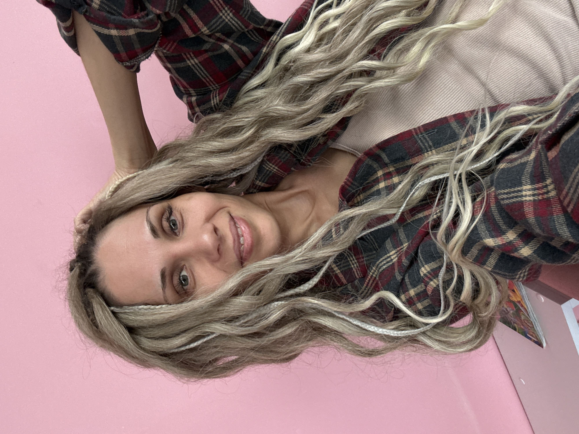 A woman with blonde wavy hair and a plaid shirt is shown against a pink background. The woman is smiling and looking at the camera. The plaid shirt is a mix of red, black, and tan colors. No evidence suggests the image was taken in Rusanovo, Russia.
