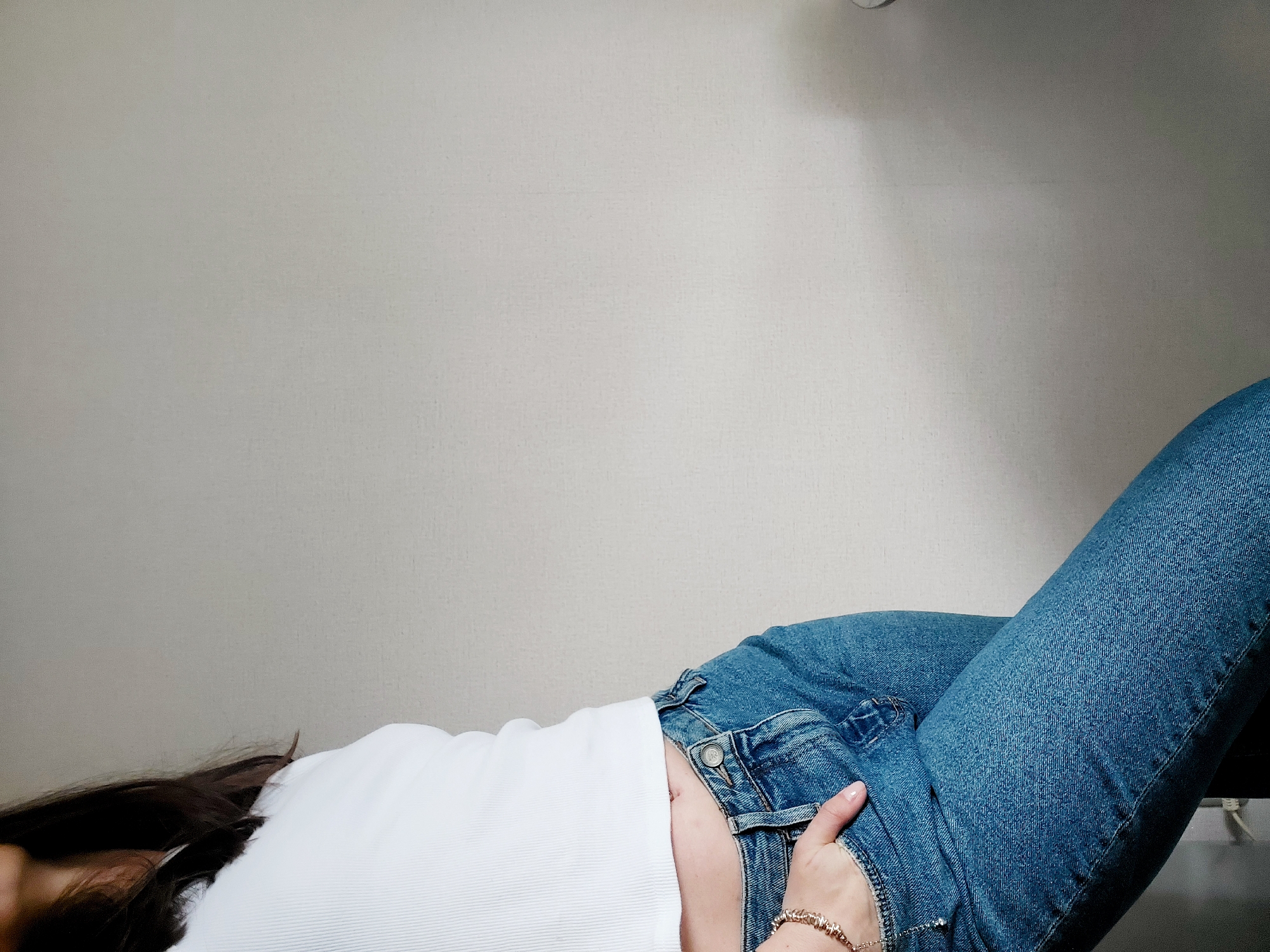 The image presents a close-up, diagonal view of a person's torso and legs, positioned against a plain, light-colored wall. The individual is wearing a white ribbed short-sleeved top and blue denim jeans.
Their body is oriented from the lower left to the upper right of the frame, with long, dark hair partially visible on the bottom left. A hand, adorned with a gold-toned bracelet and featuring neatly manicured nails, is casually tucked into the front waistband of the jeans on the right side of the frame. The denim jeans show characteristic stitching and a metal button detail on the waist.
The background is a smooth, light grey or beige wall, suggesting an indoor setting. There are no visible faces, explicit activities beyond posing or resting, or any textual elements within the image itself. The time of day cannot be determined, and while the location context provided is Chaikovsky, Russia, there are no specific visual cues within the image to indicate this.