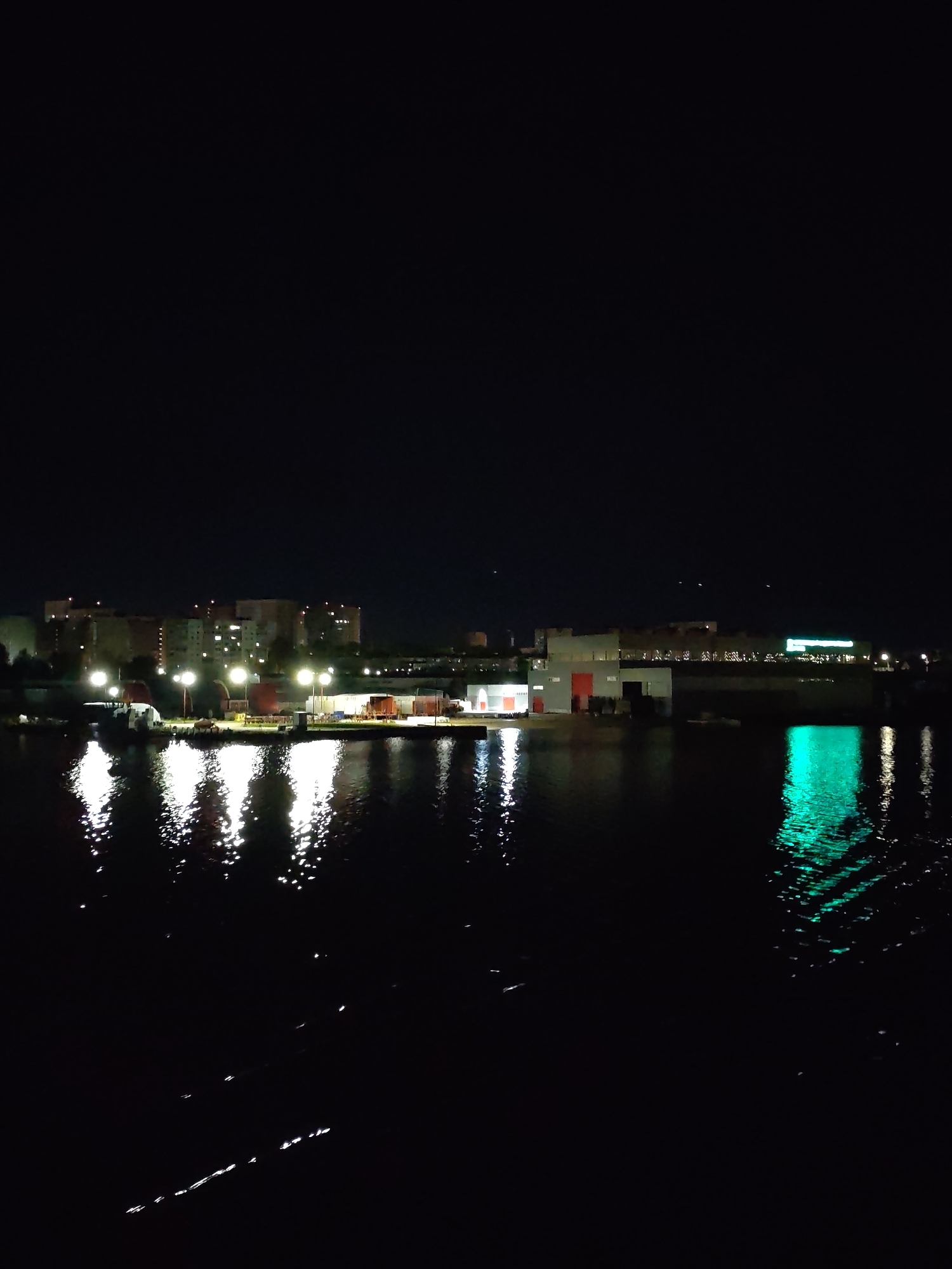 The image depicts a night scene on the Sheksna River in Cherepovets, Russia. In the background, buildings and structures are illuminated with white and green lights, their reflections visible in the dark water. A row of streetlights casts reflections onto the water's surface. The foreground is dominated by the dark, rippled surface of the river, where faint streaks of light reflect. The sky is completely dark.