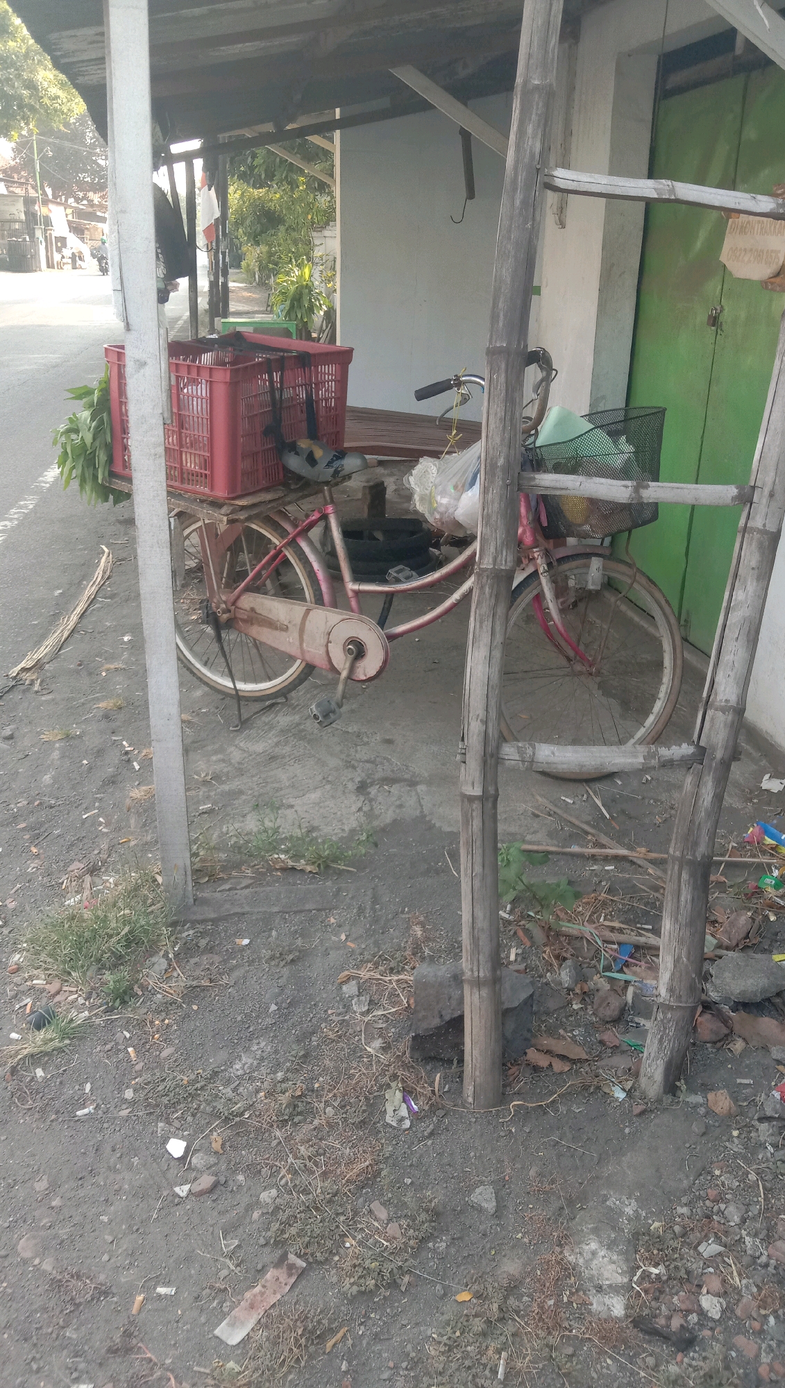 A weathered pink bicycle with a red crate containing green foliage is parked under a simple wooden and corrugated metal awning. The bicycle has a basket on the front, and a variety of items, including plastic bags, are attached to it. The scene appears to be a roadside stall in a somewhat cluttered environment, with a dirt and concrete ground surface. Debris, including cigarette butts and dried leaves, is scattered around. In the background, a white building with a green door is visible. The lighting suggests it is daytime, possibly late afternoon or early morning due to the low angle of the sun. A street is visible to the left of the awning, with a few distant buildings and trees. The location is identified as Karang Anyar, Indonesia.
