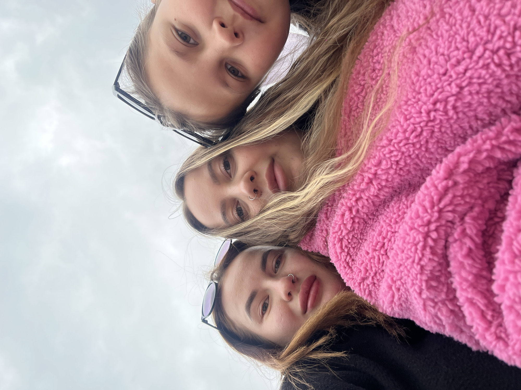 A low-angle selfie captures three young women, likely teenagers or young adults, clustered together and looking down towards the camera. The image is rotated diagonally, showing their faces from chin-up against a bright, overcast sky.
The topmost individual has light brown hair and eyeglasses resting on her head. The middle subject has blonde hair, a nose ring (hoop), and is wearing a prominent bright pink, fluffy, sherpa-like jacket. The woman at the bottom has brown hair, sunglasses perched on her head, and also a nose ring (hoop); she is wearing a dark-colored jacket, likely black.
Their expressions are generally neutral or faintly smiling, indicating a relaxed posture for the photograph. The background consists solely of a uniformly gray, overcast sky, suggesting an outdoor setting during daytime with no direct sunlight. The scene is set in Novopokrovskaya, Russia. There is no visible text in the image.
