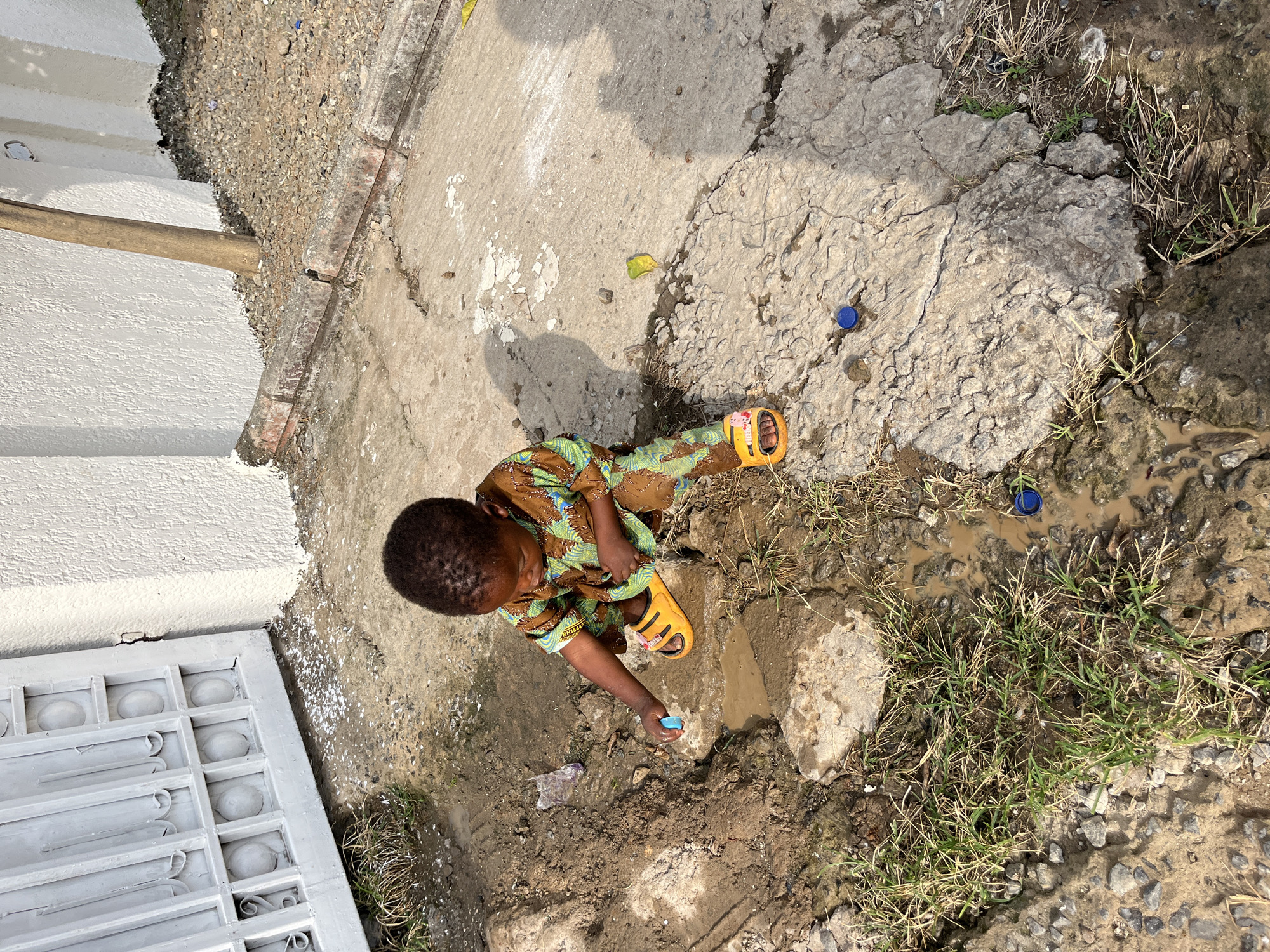 A young child, presumably a boy, is playing in a puddle of dirty water in an urban environment. The child is dark-skinned, dressed in a brown, green, and yellow patterned shirt and wearing yellow sandals. He is squatting near the water, holding a blue object in his right hand. The ground around him is a mixture of cracked concrete, dirt, mud, and small patches of grass. There are also blue bottle caps scattered near the puddle. The environment appears to be outdoors near the edge of a building or structure, with a gray door or wall visible to the left. The image was most likely taken in daylight in Abuja, Nigeria.