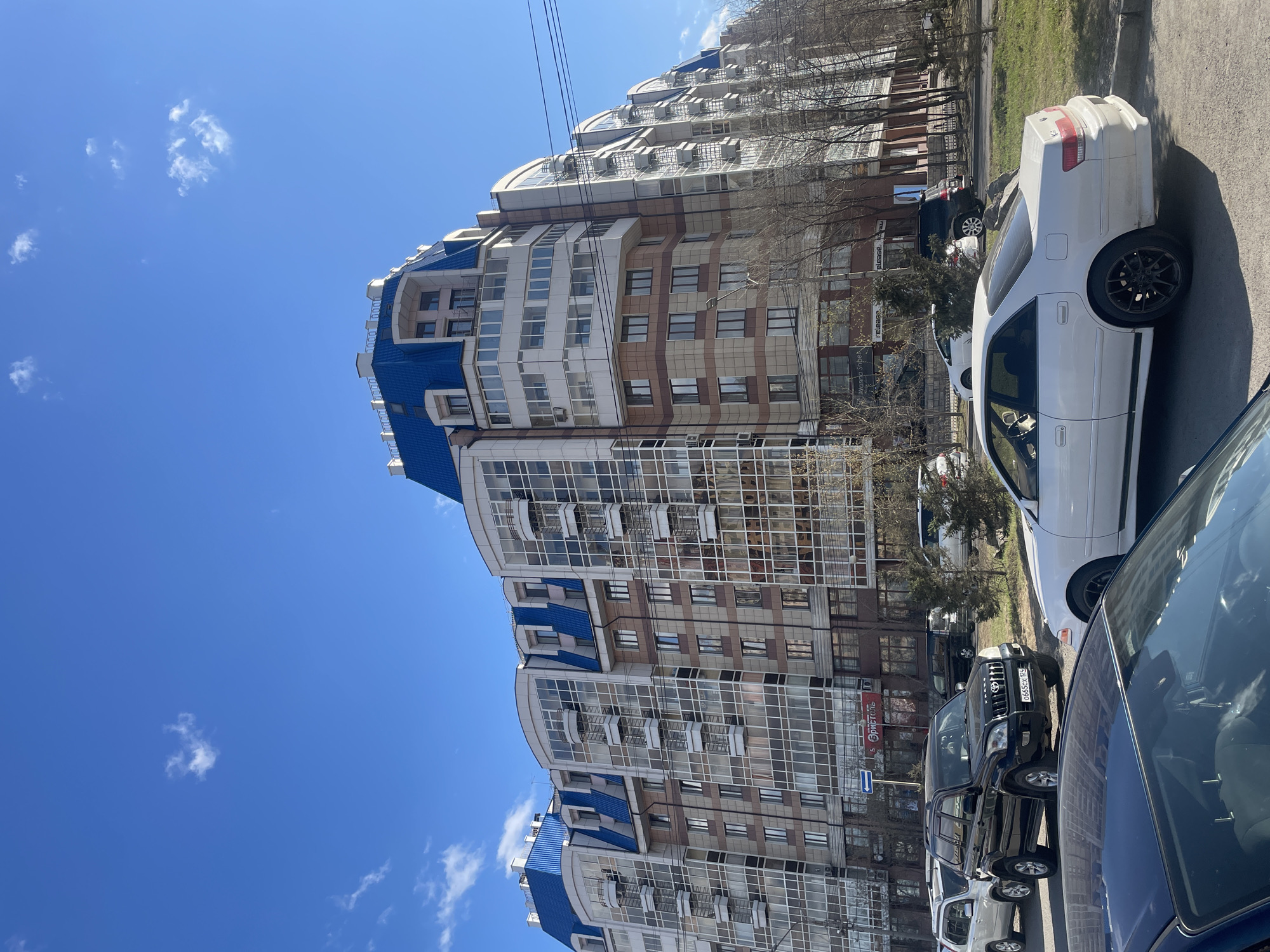 The image depicts a street scene in Krasnoyarsk, Russia. A tall, modern apartment building with a distinctive blue roof and glass-paneled facade dominates the left side of the frame. The building has a stepped design, with multiple levels. Parked cars line the street along the building's base, including a black SUV with the license plate "O665CX 124". The sky is clear and blue with scattered white clouds. Some trees are visible between the building and the parked cars.