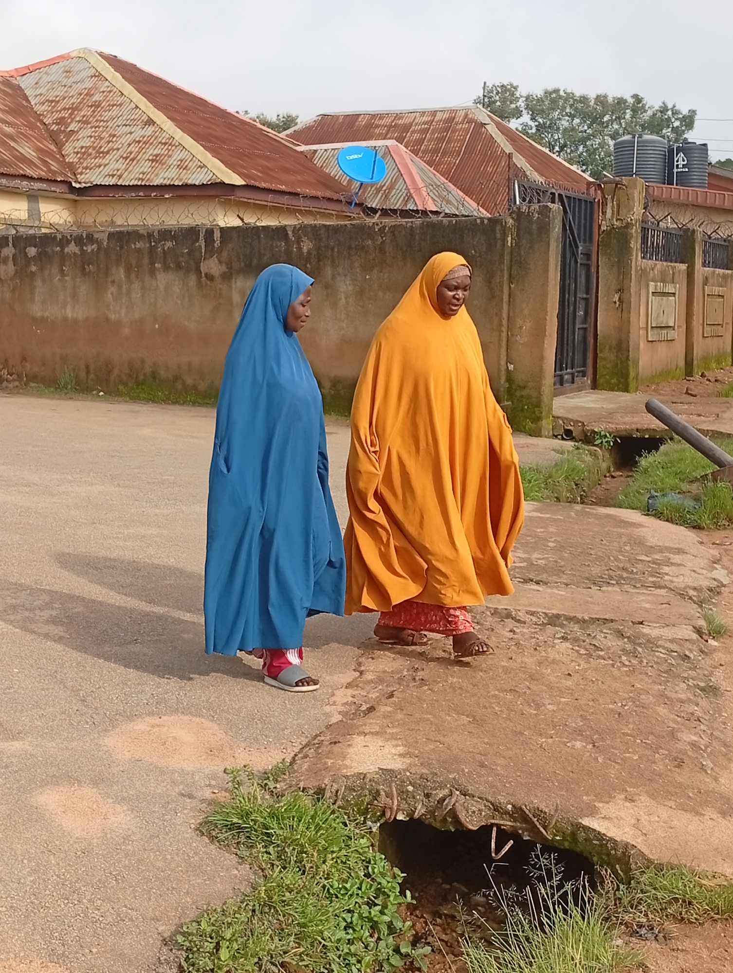 The image shows two women walking on a paved road in Jos, Nigeria. The woman on the left is wearing a blue hijab and a blue dress. The woman on the right is wearing an orange hijab and an orange dress with a red and white patterned skirt and sandals. Behind the women, there is a beige wall topped with barbed wire, and two corrugated iron roofs of houses. There are also two large black water tanks on top of a building in the background. The road is cracked and uneven, with some patches of grass growing along the edges. There is a drainage culvert in the foreground with some metal rebar sticking out. The sky is overcast.