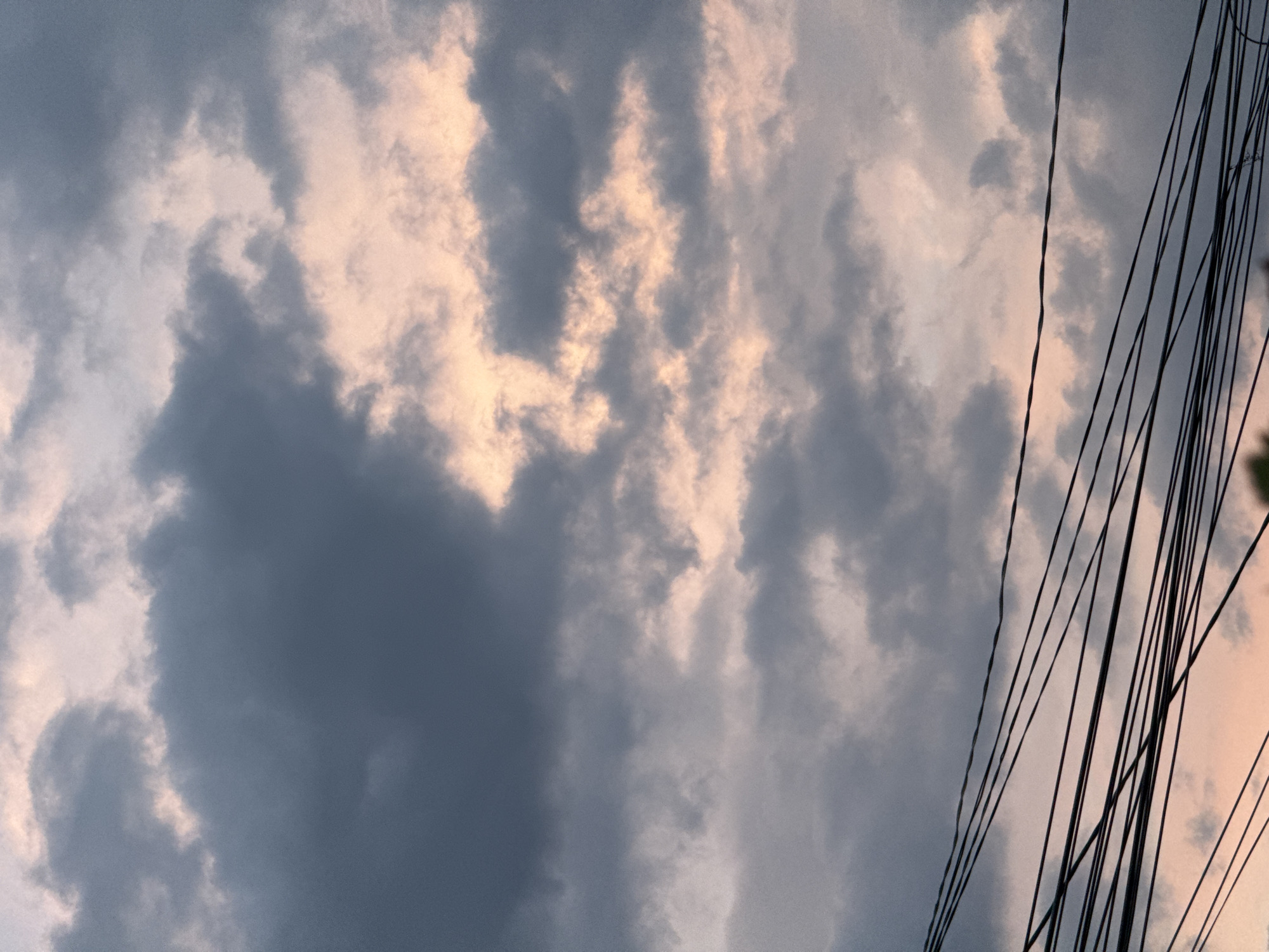This is a low-angle, outdoor shot of a cloudy sky with a tangle of power lines on the right side. The clouds are a mix of dark gray and lighter, pinkish hues, suggesting either sunset or sunrise, or perhaps stormy weather. The power lines are thin and black, with some appearing twisted or frayed. They are tightly clustered and run diagonally across the right portion of the frame, obscuring some of the sky. The overall mood is somewhat dramatic and possibly melancholic due to the dark clouds and the presence of the intrusive power lines. There are no people or specific landmarks visible to indicate the exact location beyond the provided context of Mikhaylovsk, Russia. The image is composed in portrait orientation.
