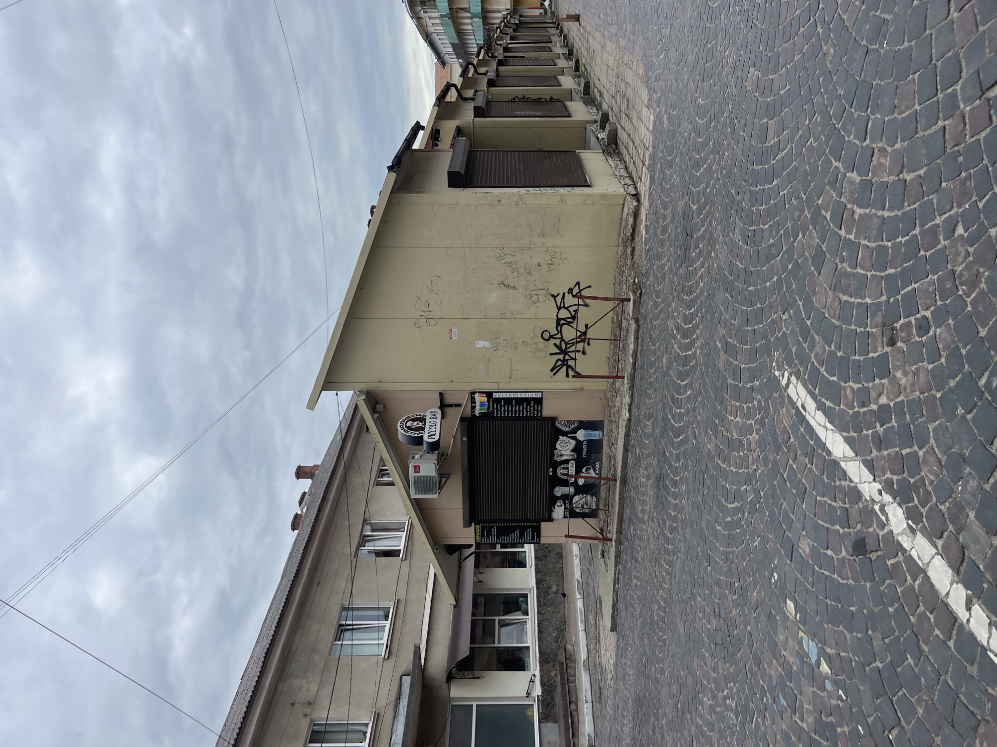 The image shows a street scene in Drohobych, Ukraine, on a cloudy day. In the foreground, a cobblestone street is visible with a white line painted on it. To the right, a row of small, tan-colored kiosks with closed shutters lines the street. The first kiosk has graffiti on its wall and a sign that reads "PICCOLO BAR". A menu with pictures of drinks and food is posted on its side. To the left of the kiosks, a larger building with windows and a gray roof is partially visible. Another building with balconies is seen in the background on the right. The overall impression is a commercial area with several small businesses.