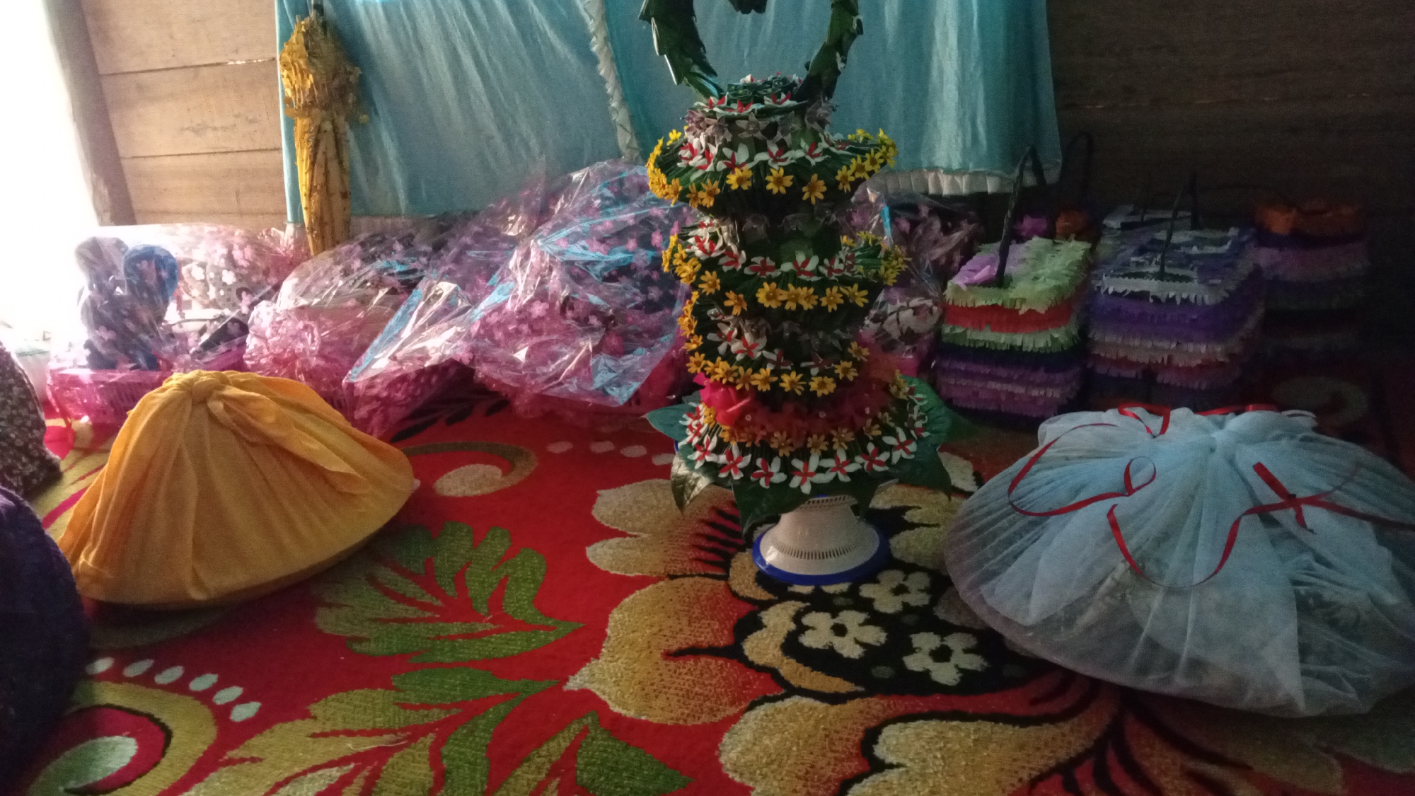 The image depicts an indoor scene with several objects on a red, floral patterned carpet. In the center, a tall, ornate decoration made of green leaves and small yellow and white flowers stands on a white base. To the left of this centerpiece, there are several bundles wrapped in clear plastic with pink or purple paper. A bright yellow fabric-covered object is also visible on the carpet. To the right of the centerpiece, there are several colorful, rectangular objects with fringed edges, possibly piñatas, in shades of purple, green, and orange. Further to the right, a large, round object is covered in white sheer fabric, tied with red ribbons. In the background, a light blue curtain hangs, and wooden planks are visible on the left. The scene appears to be a preparation for an event or a display of gifts. The location is Meunasahdayah, Indonesia.