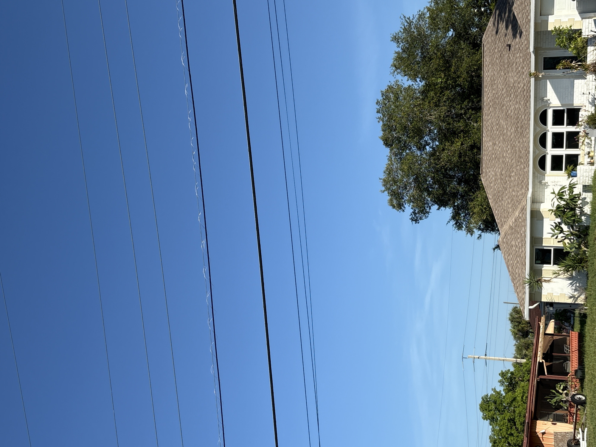 In the city of North Port, United States of America, this image captures a residential scene under a clear blue sky. The foreground features a white house with a brown shingled roof, characterized by arched windows. To the left of the house is a smaller structure with a reddish-brown roof and orange siding, appearing to be a carport or shed, with a trailer visible underneath. Dense green trees form a backdrop behind these structures. Several power lines are visible crisscrossing the sky, with some exhibiting a wavy pattern, suggesting they might be supporting small, reflective ornaments or insulation. The overall impression is a typical suburban landscape bathed in bright daylight.