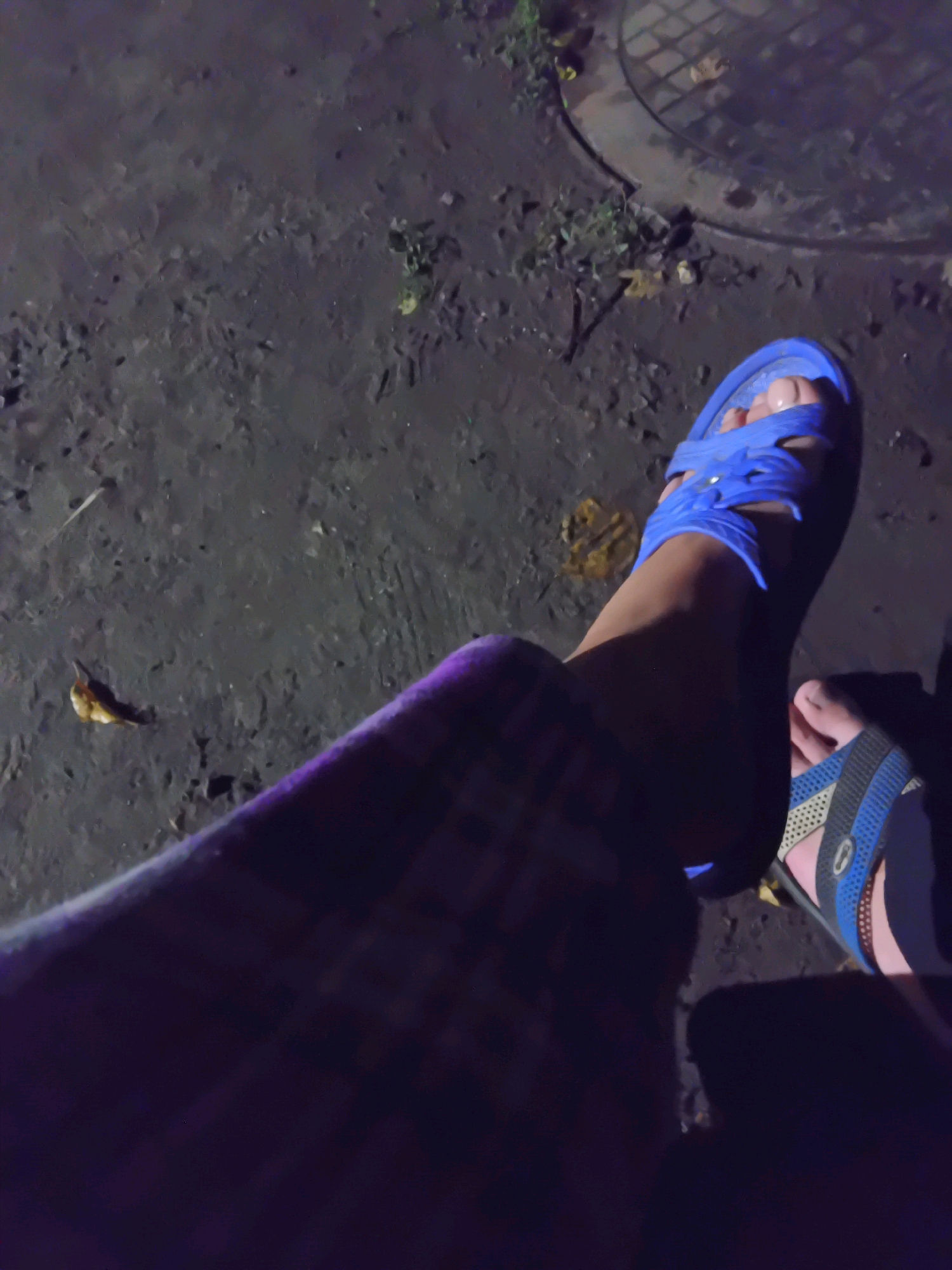 This image shows a person's legs and feet, wearing blue and blue-and-grey flip-flops, positioned on a dark, wet, uneven ground surface with some scattered leaves and debris. A circular metal manhole cover is visible in the upper right background, along with some vegetation. The lighting suggests it is nighttime or a very dimly lit area. The scene appears to be an outdoor urban environment, potentially a street or pathway in Chuhuiv, Ukraine.