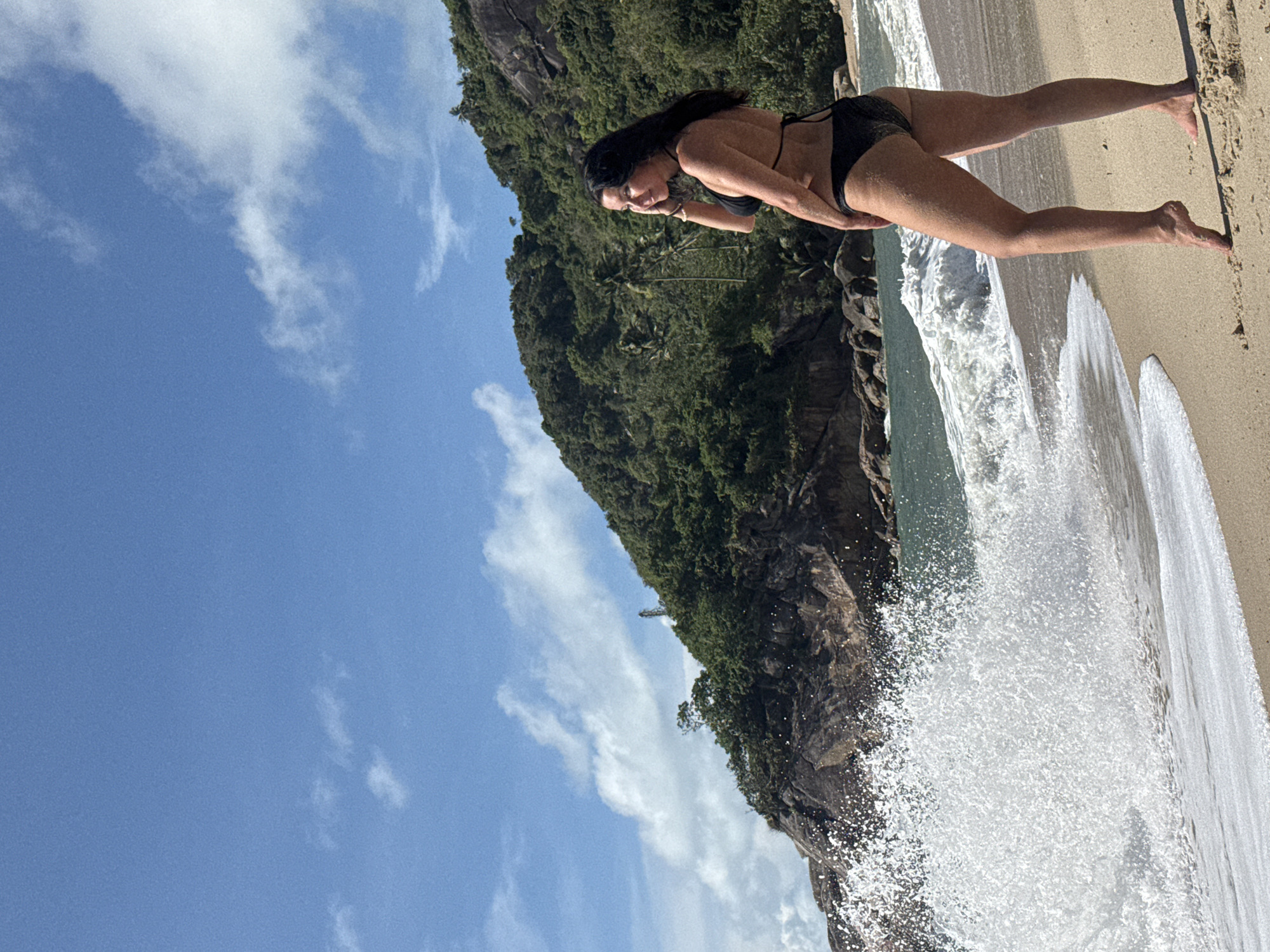 A woman is in a black bikini, standing on a sandy beach in Baan Thong Nang, Thailand. A rocky, tree-covered cliff rises behind her. Waves are crashing around her feet. The sky is blue with scattered white clouds.
