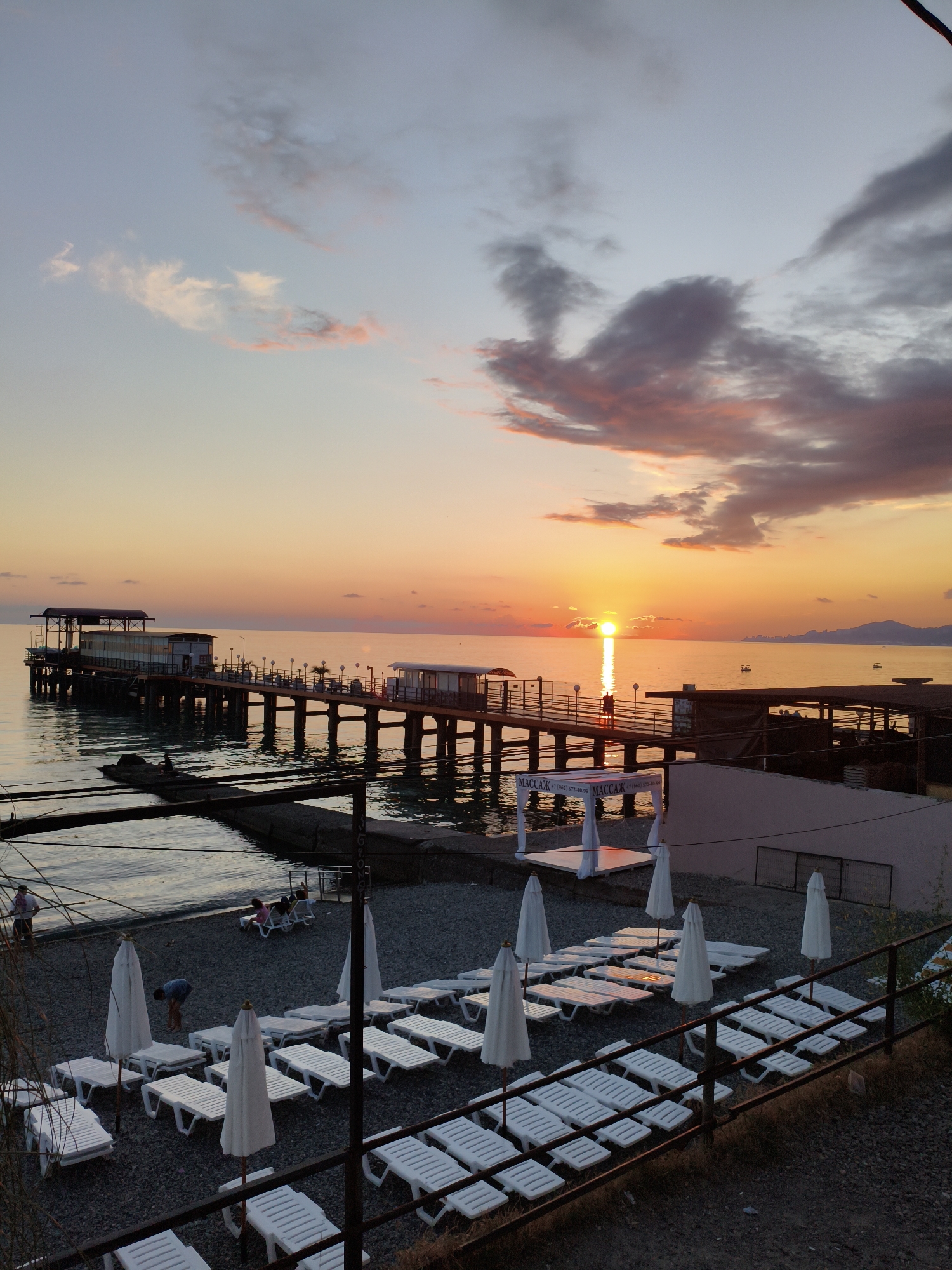 This is a photograph taken at sunset on a pebble beach in Sochi, Russia. A pier extends into the calm sea, with a building at the end. The sky is a vibrant orange and pink, with clouds scattered across it. In the foreground, rows of white lounge chairs and closed umbrellas are arranged on the beach. A few people are visible on the pier and on the beach, appearing to be enjoying the sunset. The water is dark and reflects the colors of the sky. The overall impression is one of a peaceful evening at the seaside.