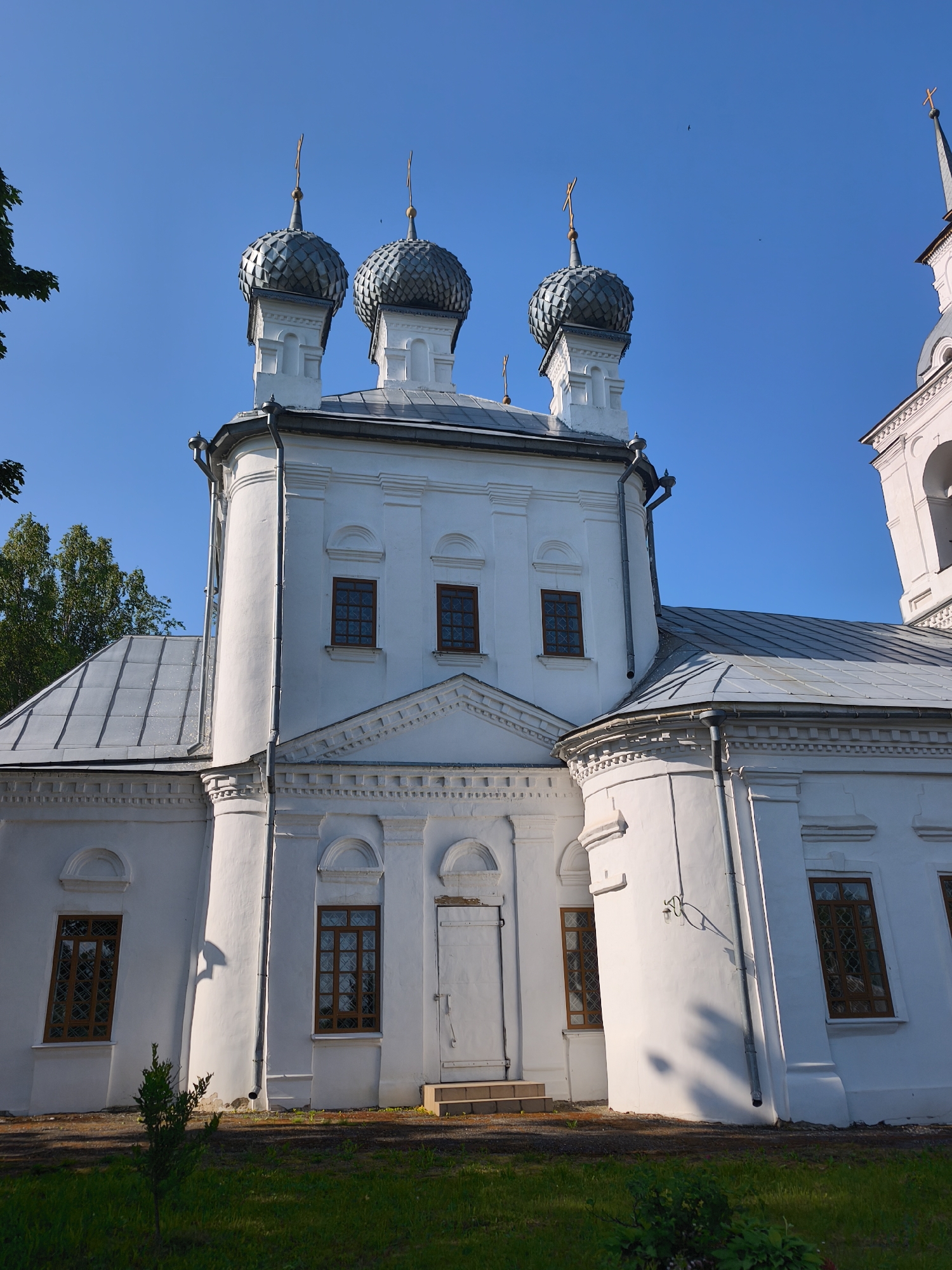 The image shows the exterior of the Church of the Nativity of the Most Holy Theotokos in Kady, Russia. The white stone church has multiple domes with crosses on top, and a grey sloped roof. The church features several windows with brown frames and intricate patterns, as well as a white door with a small set of steps leading up to it. The building is surrounded by green grass and a few trees are visible in the background. The sky is clear and blue.