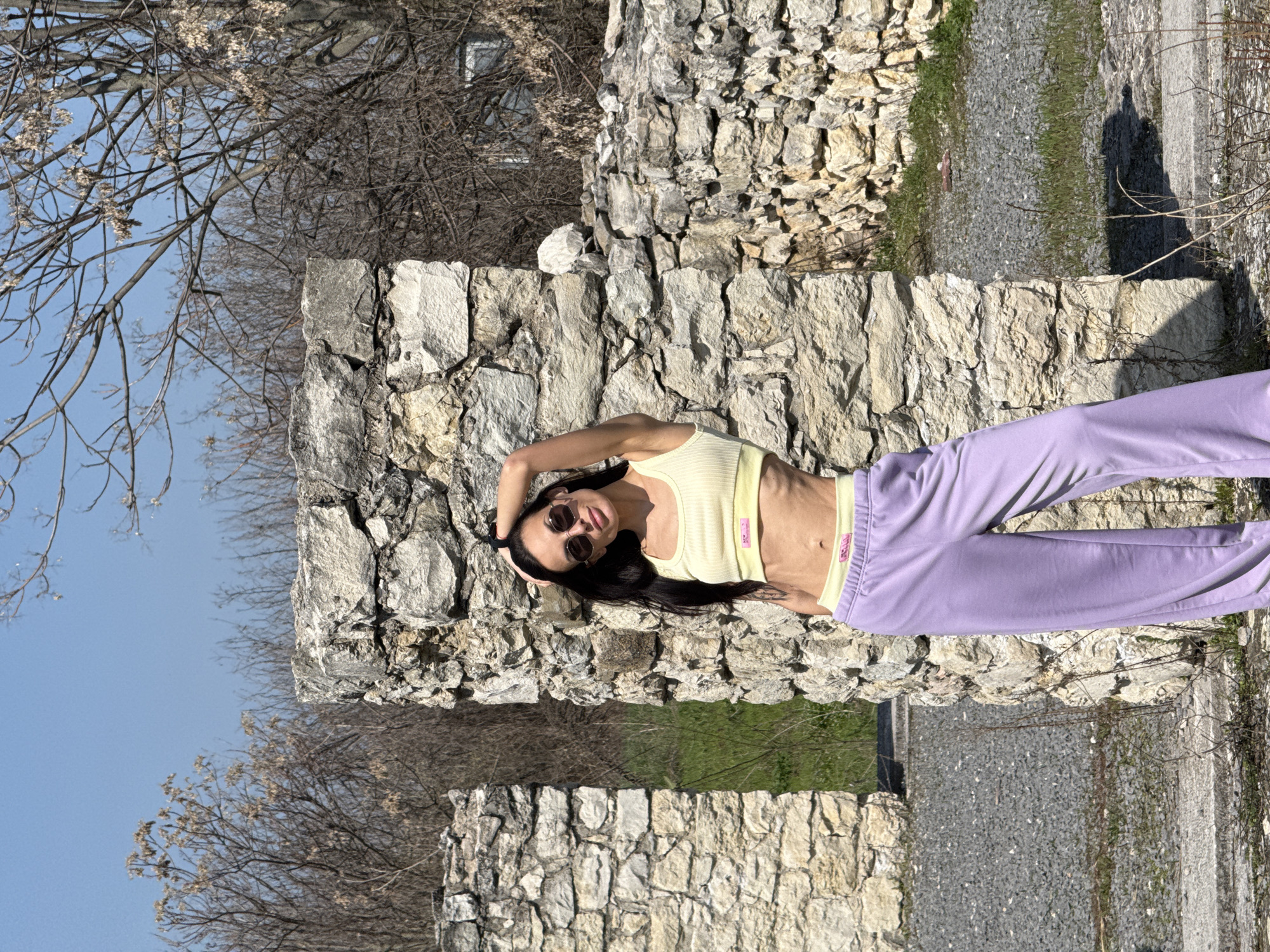 A woman is lying on a stone structure in a seemingly outdoor setting. The background shows trees and what appears to be sky. The image is oriented in such a way that the scene looks sideways, with the ground appearing to be vertical. The woman is wearing a yellow top and purple pants, and she is wearing sunglasses. The structure looks like an old stone wall. It is located in the Kaylaka Park, Pleven, Bulgaria.