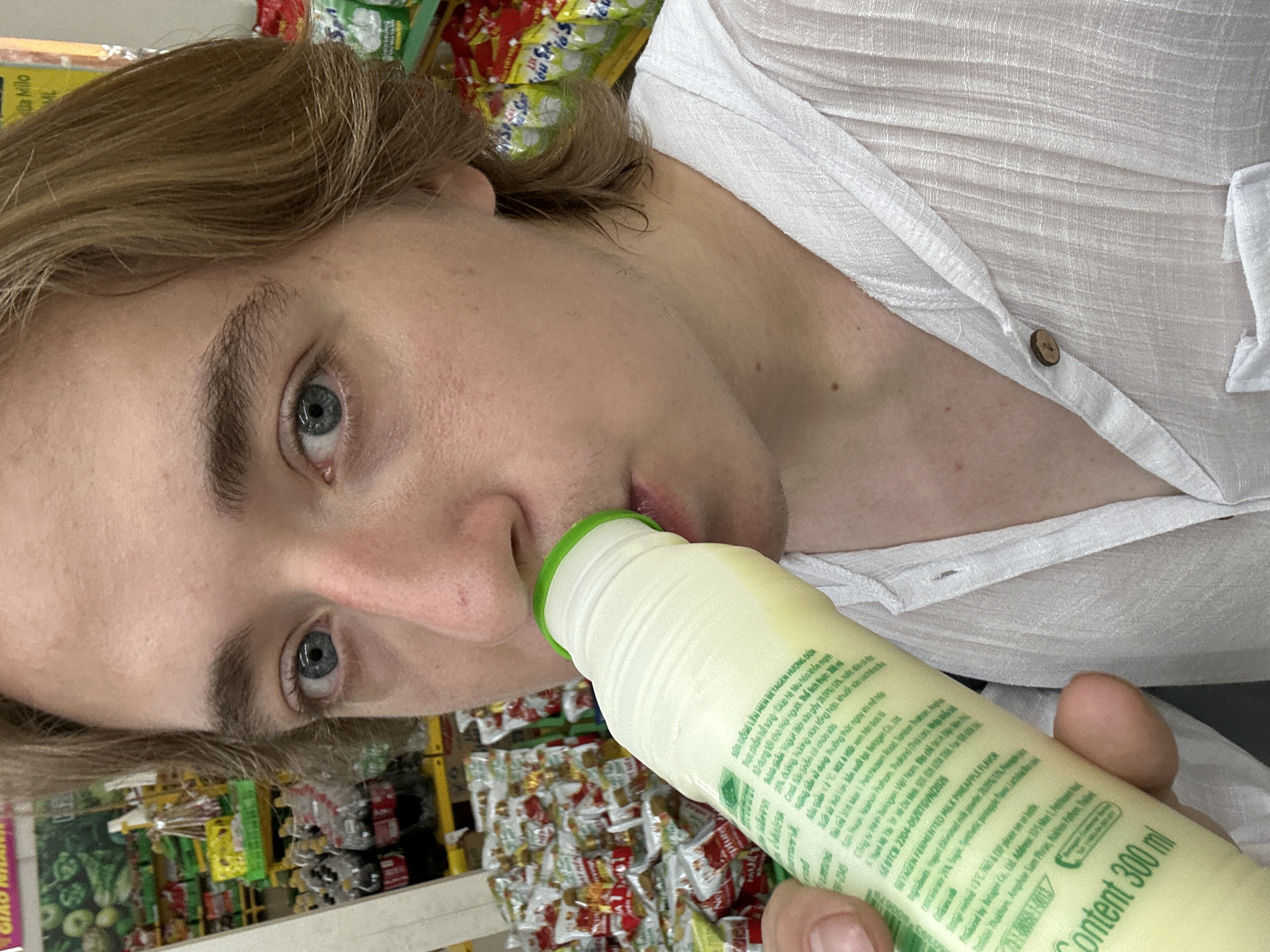 In An Hữu, Vietnam, a close-up shot captures a person drinking from a bottle of fermented milk. The person, with light skin and blonde hair, is wearing a white, textured shirt. The bottle, primarily light green with white and green lettering, reads "Content 300 ml". In the background, shelves are stocked with various food items and drinks, suggesting a retail environment such as a supermarket or convenience store. The lighting appears to be artificial, typical of indoor lighting.