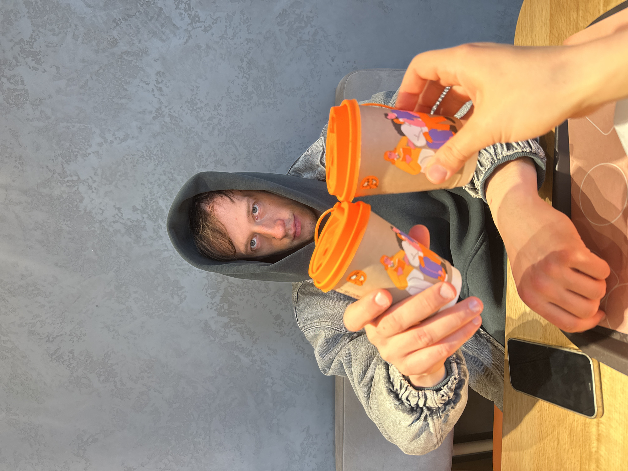 An indoor scene is captured from a slightly elevated, angled perspective, showing two individuals engaged in a casual interaction. In the left portion of the image, a man is visible from the chest up, wearing a dark gray hooded sweatshirt over a light-colored denim jacket. He has short brown hair and looks directly at the camera with a neutral expression.
On the right, another person's hands are extended towards the man, holding two disposable coffee cups. These cups are beige with vibrant orange lids and feature colorful, cartoon-style illustrations of what appear to be fennec foxes or similar small animals. One cup is positioned slightly above the other.
The immediate foreground on the right reveals a smooth, light wooden table or counter surface. A black smartphone, resembling an iPhone, rests flat on this surface to the right of the cups. The background on the left features a distinct, light gray wall with a textured, almost stucco or concrete finish. A portion of a gray upholstered armchair or sofa is also visible behind the man.
The overall lighting is bright and even, indicating a daytime setting indoors. The activity suggests one person is offering or handing drinks to the other, creating a friendly and informal atmosphere. No discernible text is visible on any objects.