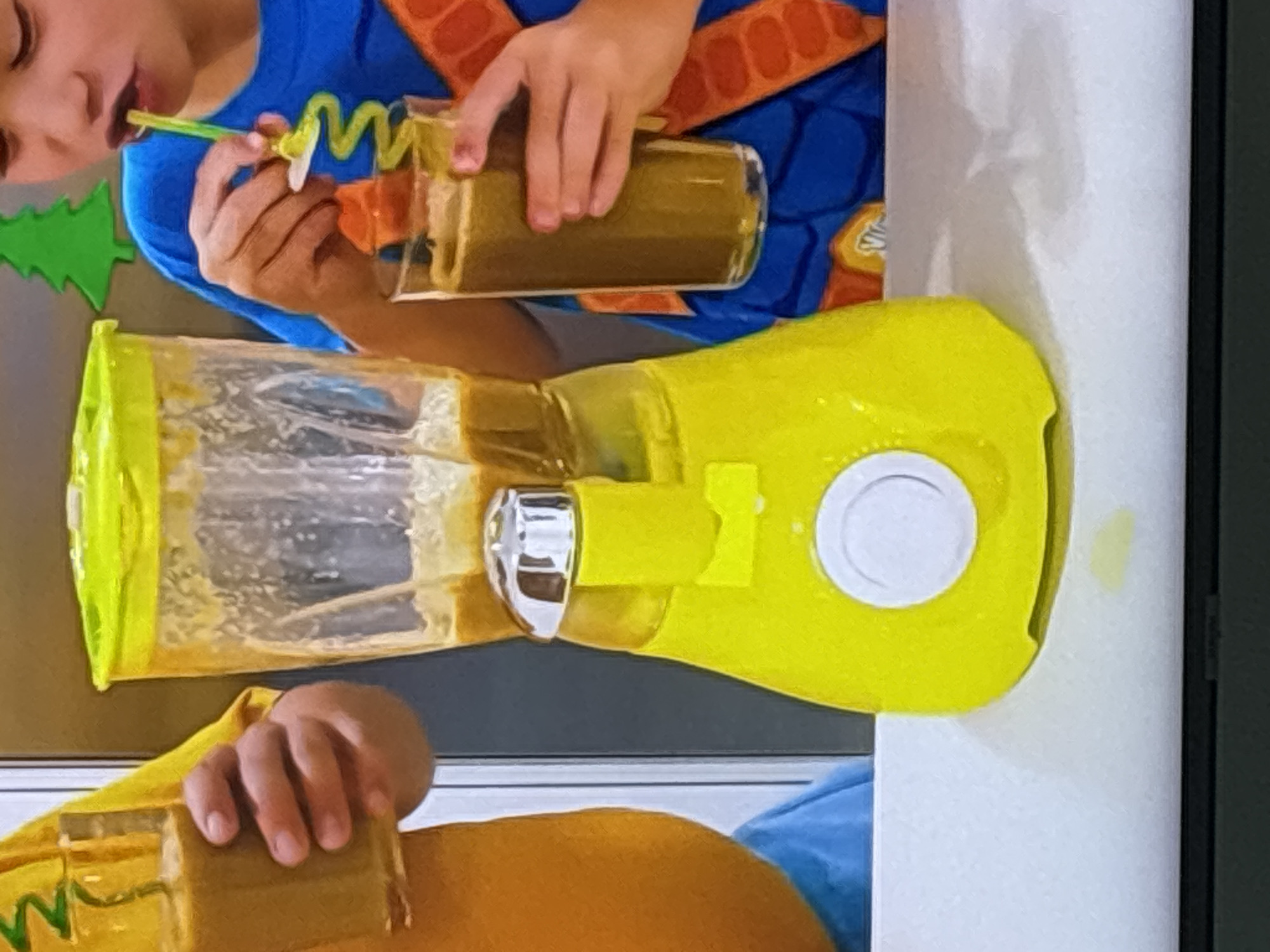 In a close-up shot, a vibrant yellow blender is positioned on a white surface. The blender's pitcher contains a light brown liquid, with some residue visible on the inside. A silver-colored lid or component is affixed to the top of the pitcher. Below the pitcher, a round, white button is prominently featured on the blender's base.
To the left of the blender, a person wearing a yellow shirt is partially visible, holding a glass filled with the same light brown liquid. Their hand is grasping the glass.
To the right of the blender, another person, wearing a blue shirt and what appears to be an orange costume with some decorative elements, is holding a glass of the brown liquid. They are actively drinking from the glass using a coiled, green straw. The background features a blurred green, Christmas tree-like cutout.
The scene appears to be indoors, likely a kitchen or a space set up for preparing beverages. There is no discernible information to suggest the location is in Dubai, United Arab Emirates, or any specific city. The lighting is bright and appears to be coming from overhead.