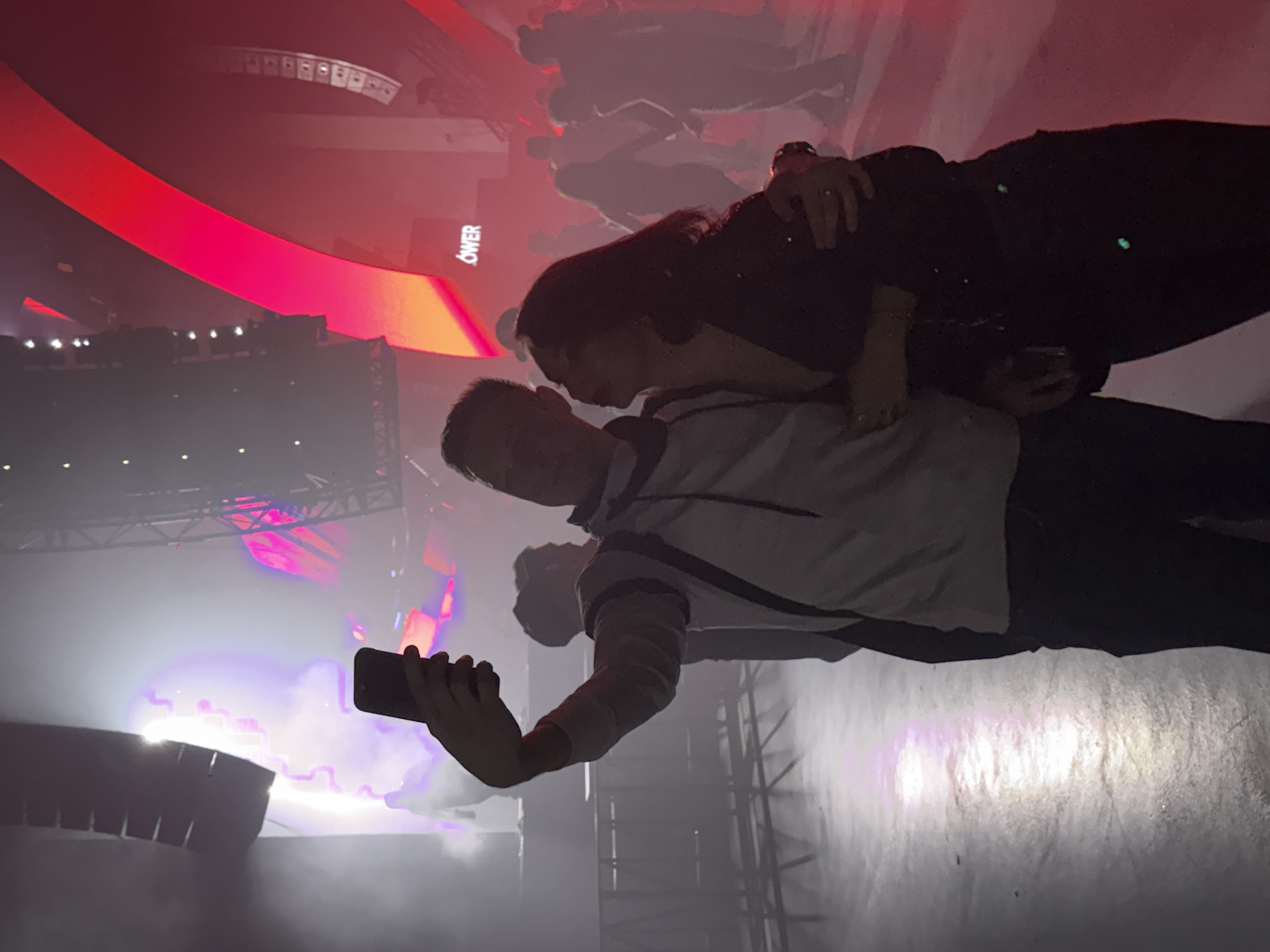 The image captures an intimate moment between two individuals at what appears to be a vibrant indoor concert, club, or festival. The scene is illuminated by dramatic stage lighting, predominantly in shades of red, pink, and purple, with strong light sources creating haze effects.
In the foreground, a man and a woman are embracing. The woman, with long dark hair, leans her head close to the man's, her arm wrapped around his shoulder/neck. She wears a dark top. The man, wearing a light-colored top with dark stripes or accents, holds a black smartphone in his left hand, pointing it towards the stage or performance area, suggesting he is recording or taking a photo. Their posture indicates affection and shared enjoyment of the event.
The background reveals typical concert elements: silhouettes of other attendees forming a crowd, large stage speakers, and complex truss or scaffolding structures supporting lighting equipment. Bright beams of light cut through the smoky or hazy air, adding to the dynamic atmosphere. A prominent feature in the background is a large, curved red and orange structure, possibly a screen or decorative light panel. The fragmented word "OWER" is visible on this structure, potentially part of a larger word like "POWER". The floor beneath the subjects appears somewhat reflective and dimly lit.
The time of day is clearly night, or it's an indoor event designed to simulate nighttime. The mood is energetic and lively due to the event, yet intimate for the couple in the foreground. There are no explicit location cues within the image to confirm Khimki, Russia, other than the overall appearance of a modern event venue.