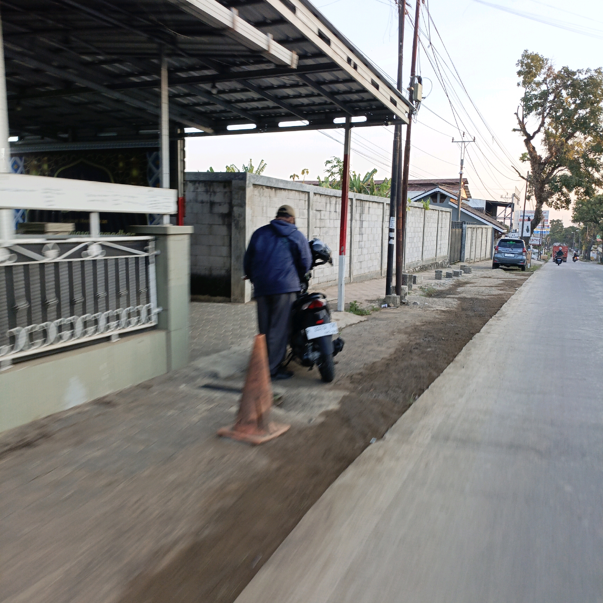 A rear view of a man wearing a blue jacket and a tan cap, standing next to a black motorcycle. The motorcycle is parked on a dirt path next to a paved sidewalk. An orange traffic cone is placed in front of the motorcycle. In the background, there is a long concrete wall, several trees, power lines, and buildings. The scene appears to be along a street in Salatiga, Indonesia, with a car and two motorcycles visible further down the road. The lighting suggests it is either early morning or late afternoon. The image is somewhat motion-blurred, indicating movement during the capture.