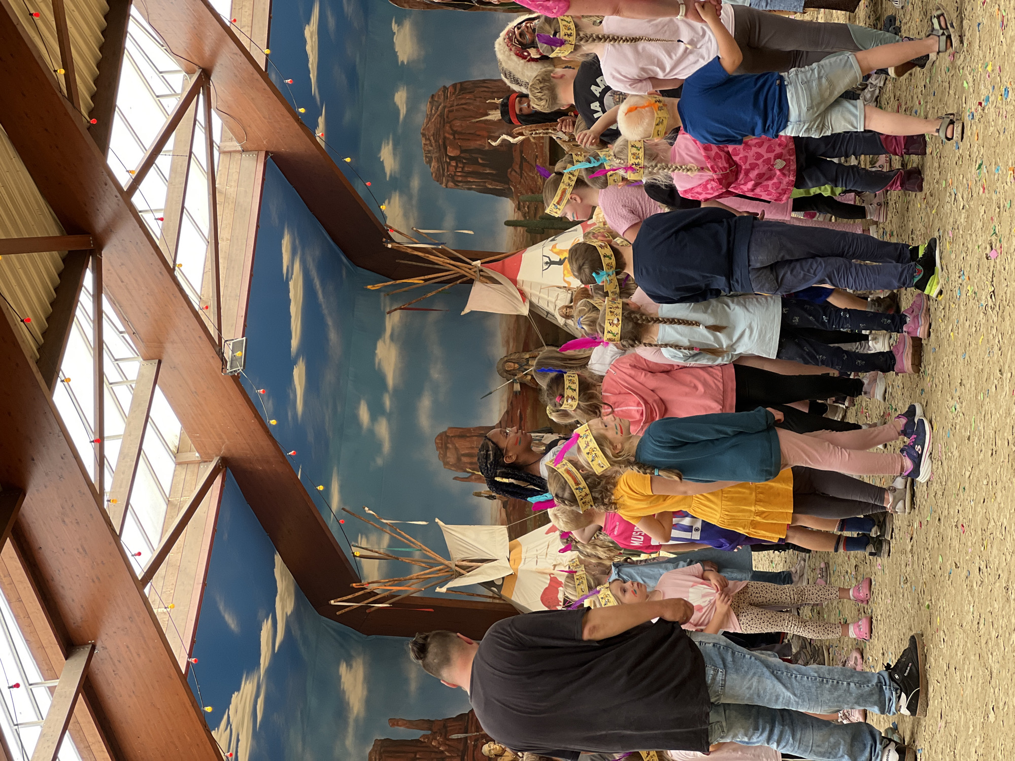 This image depicts an indoor event space decorated with a Western or Native American theme. The ceiling features exposed wooden beams and translucent panels, with colorful string lights hanging down. The backdrop is a mural of a desert landscape with mesas and a blue sky with clouds.
In the foreground, a group of children and a few adults are gathered on a sandy floor strewn with confetti. Many of the children are wearing headdresses and sashes, suggesting a themed costume party or performance. Some of the children have face paint, resembling war paint.
Several teepee structures are visible throughout the space, some appearing to be decorative props and others potentially part of the activity. One teepee in the background is adorned with colorful ribbons. A man in a feathered headdress and traditional attire is visible in the middle ground, facing away from the camera.
The overall atmosphere appears festive and engaging for children, likely part of an organized event. The location is identified as the Speelstad Oranje theme park in Collendoorn, Netherlands, which is known for its indoor play areas and family-friendly events.