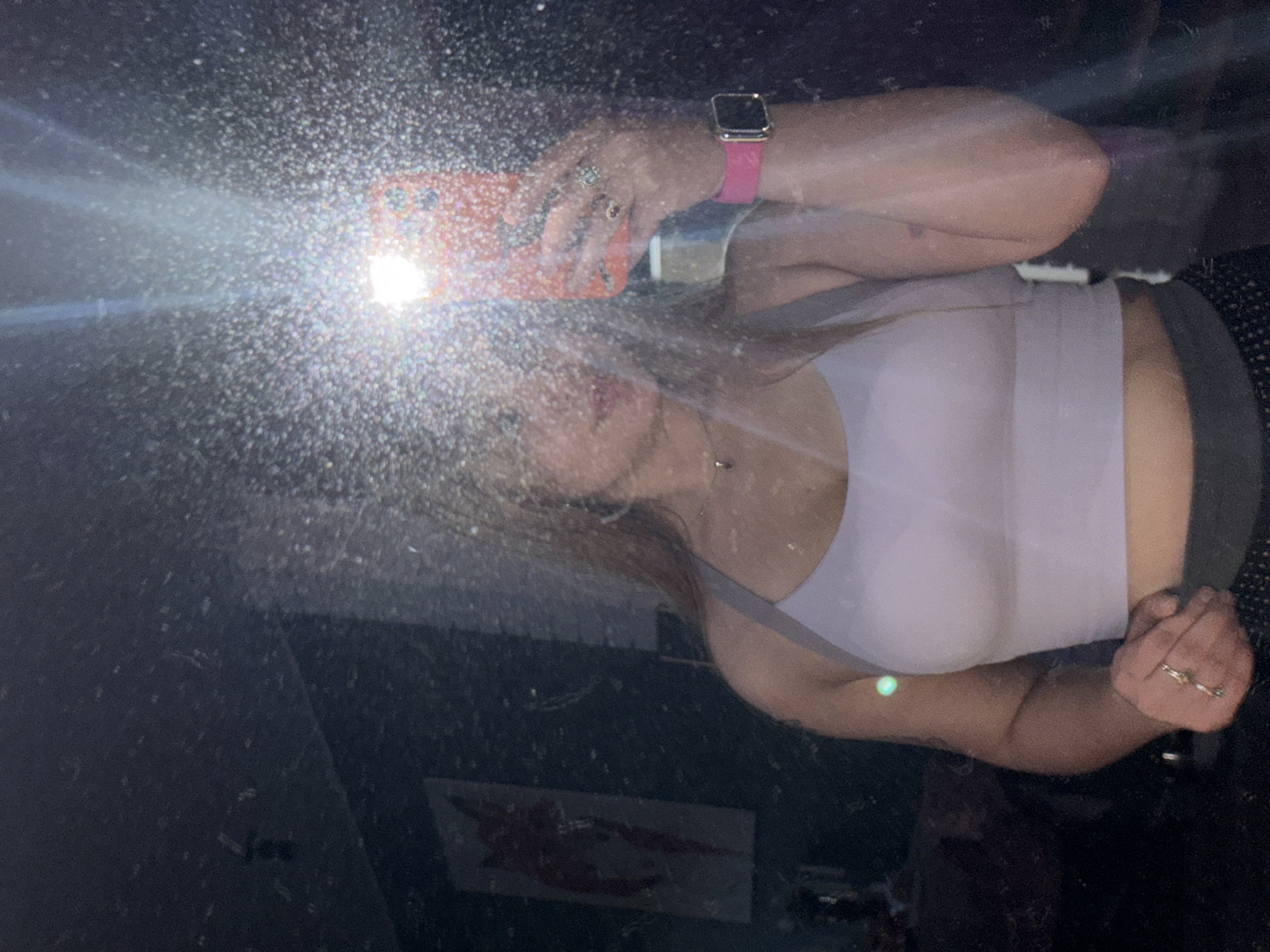 A mirrored selfie appears to be taken in Большая Шильна, Russia. A woman is holding a cellphone with a lit flash, obscuring most of her face. She is wearing a light-colored tank top, a pink watch, and several rings. Her hair is light brown. The background is dark and cluttered, with a painting or photograph of a red-colored object visible in the lower part of the mirror's reflection. The mirror is dirty and has smudges.
