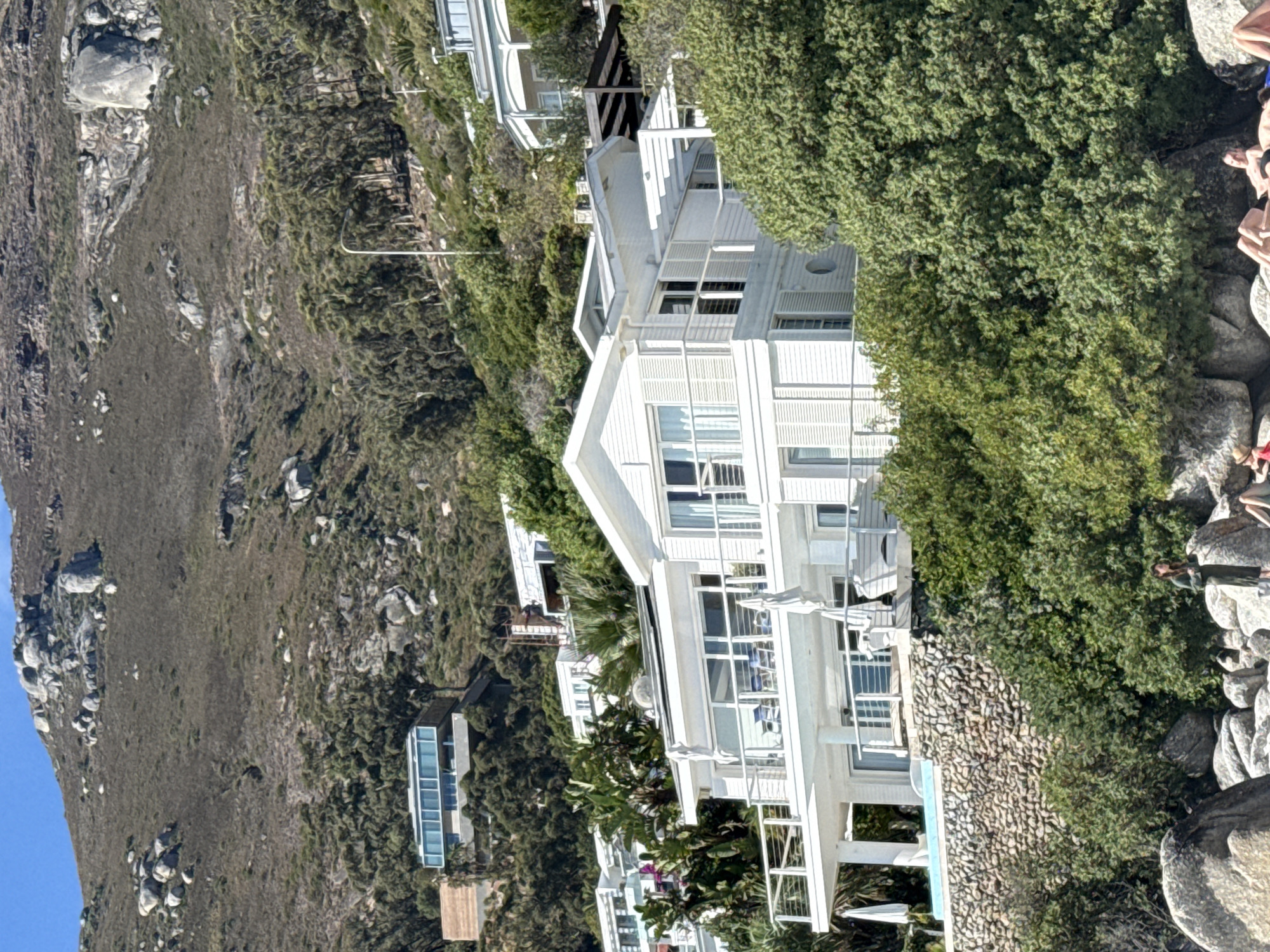 The image displays a sunlit coastal scene on a steep, vegetated hillside descending to a rocky beach, specifically located in Clifton, Cape Town, South Africa.
In the midground, a prominent multi-story white residential building with a contemporary architectural style features multiple balconies equipped with metal railings, large windows, and white louvered shutters. White patio furniture and two white patio umbrellas are visible on the upper balconies. Adjacent to the building, a small, turquoise swimming pool is present, positioned atop a robust stone retaining wall. Higher up the hillside, other white residential structures, including a distinctive modern house with extensive glass paneling, are interspersed among dense green and dry fynbos vegetation, along with numerous large grey granite boulders. A single white street light pole is visible on the upper slope.
In the immediate foreground, on a rocky beach composed of large, smooth grey boulders, one individual is standing upright, dressed in a dark, long-sleeved garment, facing towards the right side of the frame. At least three other individuals are lying prone on the boulders, indicative of sunbathing. The sky overhead is clear and blue.