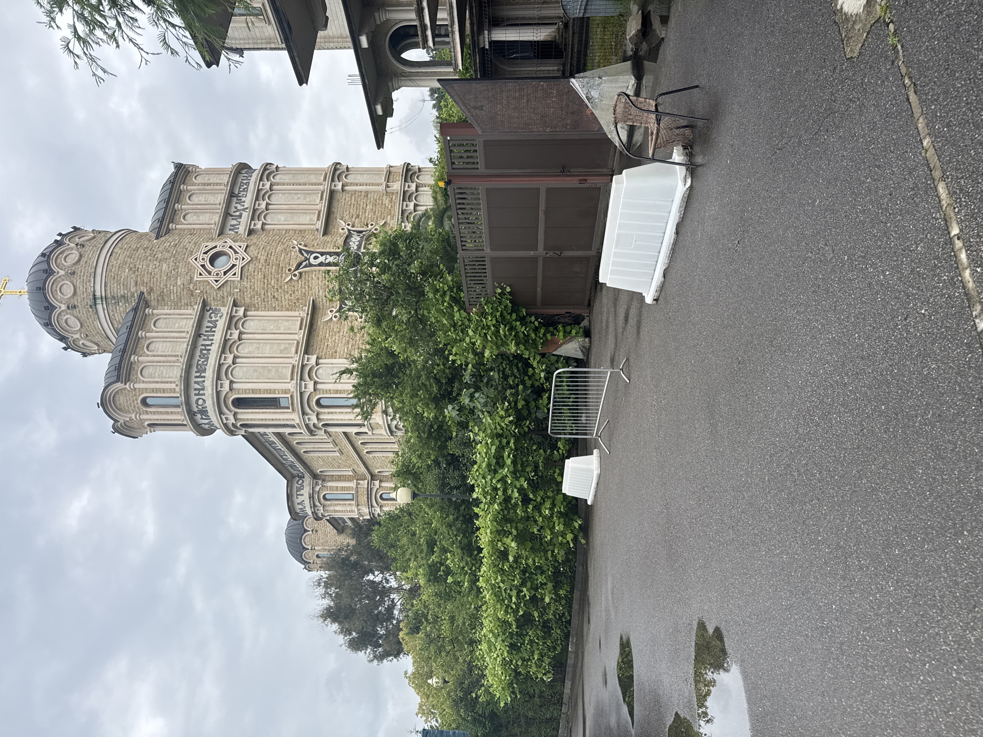 The image captures a view of the St. Michael the Archangel Cathedral in Sochi, Russia, taken from a slightly elevated position on a paved area next to the building. The cathedral, with its distinctive Byzantine Revival architecture, is a prominent feature on the left side of the frame. The grey, overcast sky indicates it is likely a cloudy or rainy day. The architecture includes a light-colored facade, rounded domes, and Cyrillic inscriptions along the exterior.
Adjacent to the cathedral's grounds is a mixture of foliage including dense green trees and shrubs that separate the building from the walkway. A silver temporary barrier and a white plastic container, both seemingly placed, are visible on the right side of the greenery. Further along the walkway, there is a brown wooden fence and gate, as well as another building with a similar architectural style to the cathedral, although less ornamented. A chair is visible, positioned adjacent to a white plastic container. The paved area shows signs of wetness with puddles indicating recent rain. The overall composition is one of a historic religious structure in a well-maintained urban setting.