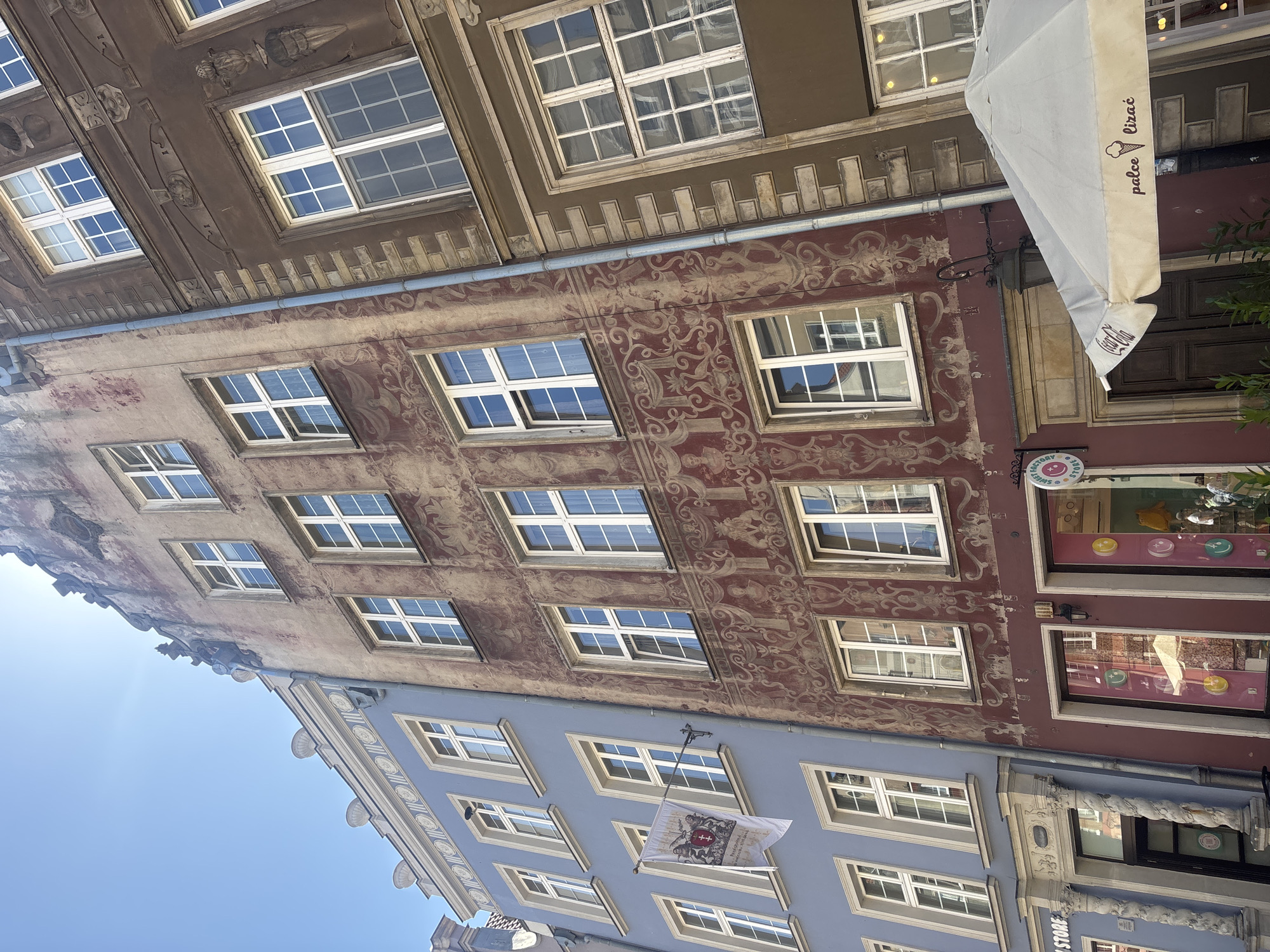 The image displays the facades of several buildings with multiple stories and windows. The buildings are situated in an urban environment, possibly a European city, given the architectural style.
The buildings feature diverse colors and decorative elements. The uppermost building has a dark brown facade with decorative moldings around the windows and on the frieze. Below it, a building with a reddish-brown facade is adorned with intricate, faded patterns in a lighter color, resembling ornate scrolls or floral designs. The windows on these upper floors are white-framed and divided into multiple panes.
The building in the foreground is painted a light blue and has a more classic facade with decorative stonework above some of the windows. Many windows are visible, all with white frames and multiple panes. A white flag with a coat of arms and text is hanging from a pole on the blue building.
An off-white awning, possibly from a cafe or shop, is partially visible on the right side of the image, extending from one of the buildings. It has text and a graphic of an ice cream cone. Below the awning, shop windows with displays can be seen.
The image is taken from a low angle, looking upwards towards the sky, which is a clear blue. The lighting suggests it is daytime, likely sunny, with shadows visible on some facades, indicating a time when the sun is not directly overhead. The overall impression is of an older, well-preserved urban streetscape.
Visible text includes "palce" (possibly part of a Polish phrase like "palce lizać" meaning "lick your fingers") and "Coca-Cola" on the awning. There is also text on a small circular sign visible in the shop window, reading "SWISS" and some other illegible words, and the flag also contains text which is difficult to read.