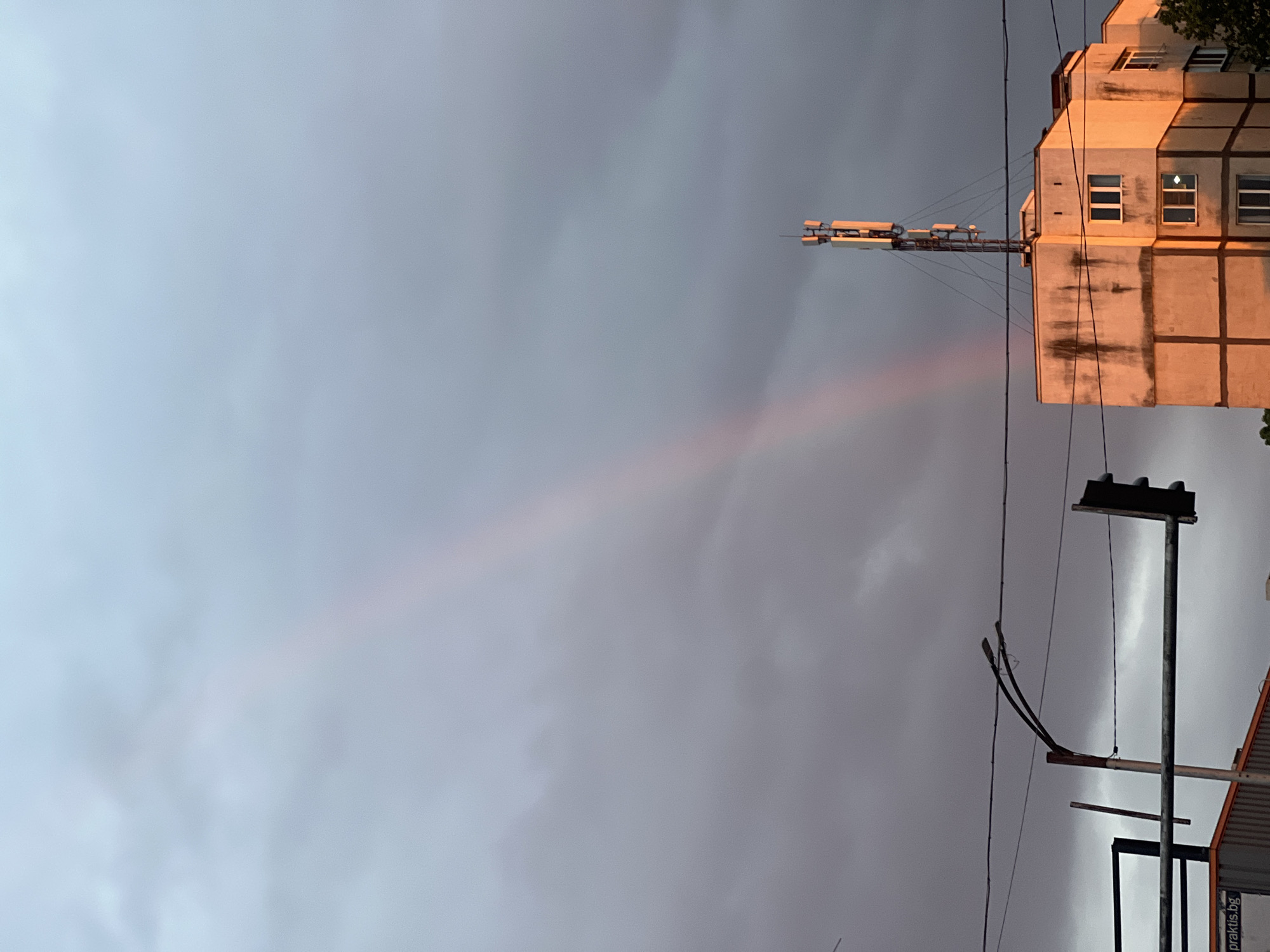 The image shows a cityscape under a cloudy sky. A partially visible rainbow stretches across the sky, left to right. In the upper right, there's a multistory building with a protruding structure that could be a communication tower. Power lines are strung across the lower portion of the frame, obscuring part of a commercial building. The location is likely Sofia, Bulgaria, based on the writing visible on the commercial building, "praktis.bg".
