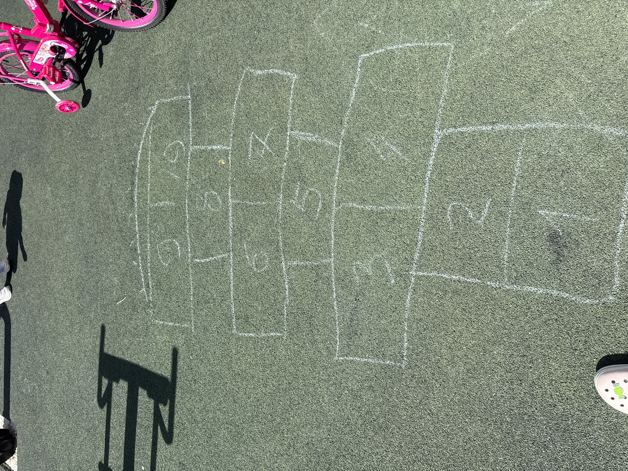 The image shows a hopscotch grid drawn with white chalk on a green, artificial turf surface. The grid consists of numbered squares and rectangles, with numbers visible including 1, 2, 3, 4, 5, 6, 7, and 9. In the background, there are shadows cast by playground equipment and two pink children's bicycles with training wheels. The scene appears to be an outdoor recreational area, likely a playground or park. The specific location in the city of Вязники, Russia is not identifiable from the image.