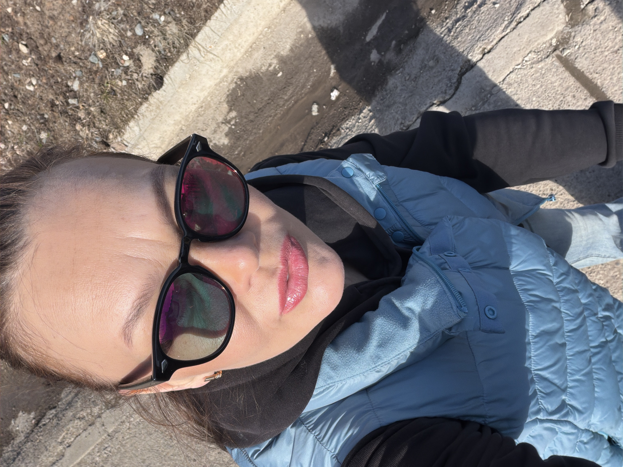 An adult female is depicted in a high-angle selfie photograph, positioned outdoors in Chaikovsky, Russia. She wears black-framed sunglasses with multi-colored reflective lenses, a black hooded sweatshirt, and a light blue quilted vest. Her lips are coated with a glossy, light pink lipstick, and a small gold-colored hoop earring is visible on her right ear. Her brown hair is pulled back. The background consists of a grey concrete surface on the left and a strip of dry, light brown earth with scattered rocks and sparse, dry vegetation on the right. A concrete curb diagonally bisects these two ground textures. A small puddle of muddy water with white debris is visible in a depression on the dirt side. Strong shadows are cast on the ground, indicating bright sunlight.
