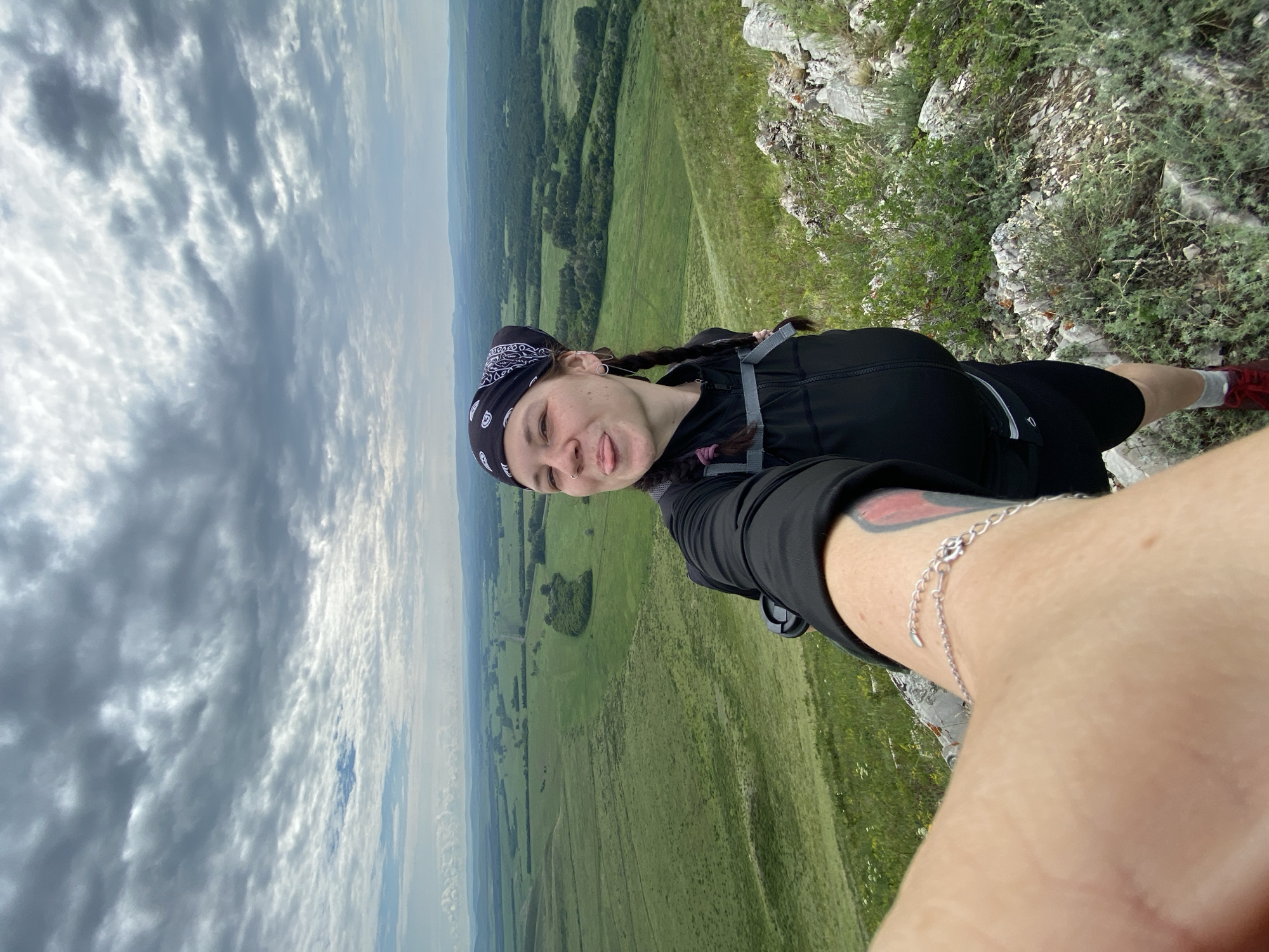 A selfie is taken by a woman standing on a rocky, grassy hilltop overlooking a vast, green valley under a cloudy sky. The woman is wearing a black bandana with a white pattern, a black zip-up jacket, and black shorts. Her dark hair is in two braids, and she is sticking her tongue out playfully. A silver bracelet is visible on her left wrist. In the background, the landscape features rolling green hills, scattered trees, and a winding road. The sky is filled with dramatic, grey clouds, suggesting an overcast day. The location is identified as Verkhnyaya Chernorechka, Russia.
