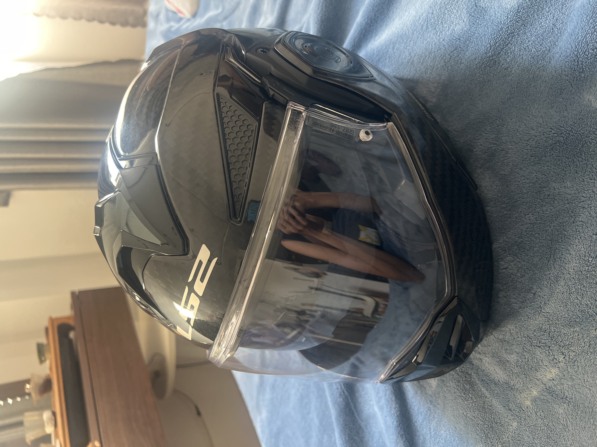 A black LS2 motorcycle helmet with a clear visor is placed on a blue, fuzzy blanket. The helmet has a carbon fiber pattern and is positioned at an angle, with its front facing slightly towards the right. The visor is partially open, revealing the interior of the helmet. The background shows a blurred image of a room with curtains and a wooden furniture piece. There is no indication that this image is located in Da Nang, Vietnam.