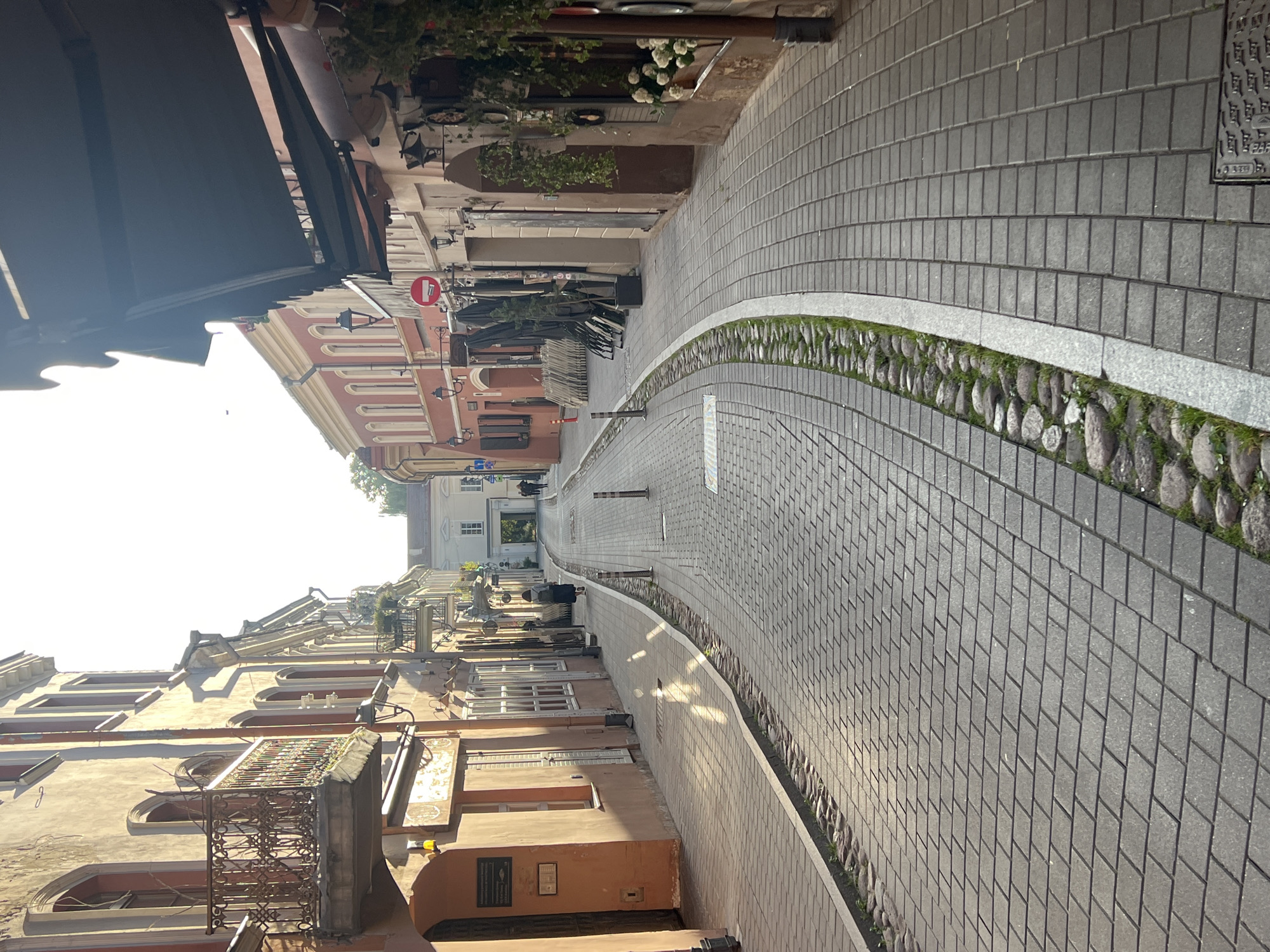 This is a high-angle shot of a narrow cobblestone street in Vilnius, Lithuania. The street is lined with historic buildings with balconies and awnings. The sun casts long shadows across the pavement, indicating it is either early morning or late afternoon. There are a few people visible walking in the distance, and outdoor seating areas for cafes are visible on the right side of the street. The street is clean and well-maintained. The architecture suggests an older European city.