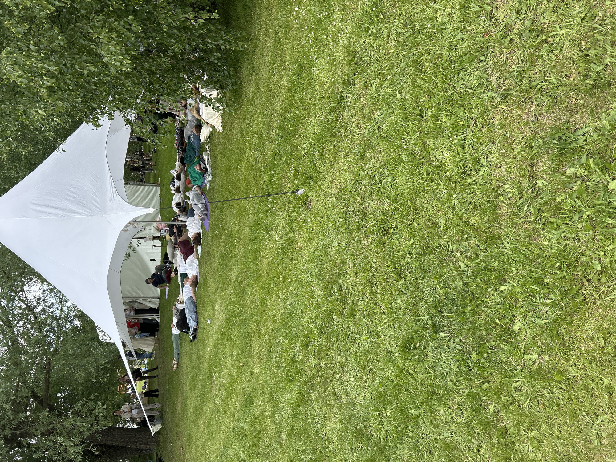 The image depicts an outdoor gathering on a grassy lawn. A large white tent is positioned on the left side of the frame, partially shaded by trees. Several people are seated and lying on the grass, extending from the tent into the open area. The lawn is green and well-maintained. A long, thin pole extends diagonally across the grass. The exact location within the city of Unknown, Unknown, is not discernible from the image.
