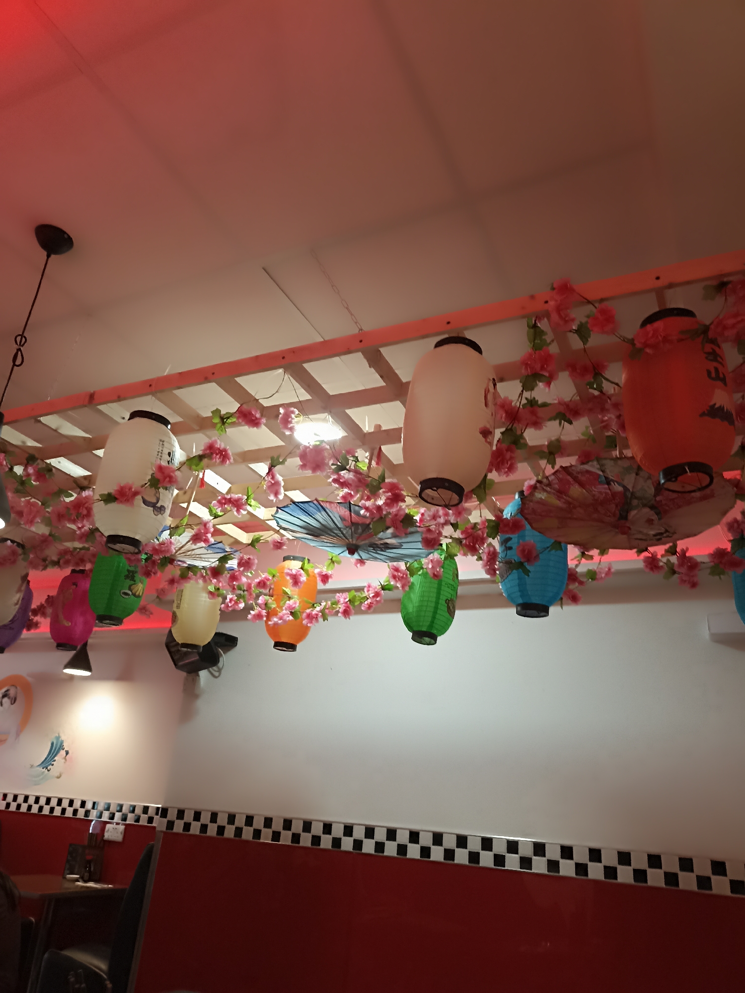 This image shows a ceiling decorated with Japanese-style lanterns and artificial cherry blossoms, suggesting an East Asian restaurant. The lanterns are of various colors and shapes, including white, orange, green, blue, and pink. Some lanterns have intricate designs on them. Several small, decorative umbrellas are also visible, interspersed with the lanterns and flowers. The ceiling itself appears to be a white grid structure, with red lighting casting a warm glow on the scene. Below the ceiling decorations, a white wall is visible, adorned with a black and white checkered border near the bottom. In the lower portion of the image, glimpses of red furniture and seating suggest the interior of a dining establishment. The scene is well-lit, with the red lighting adding to the ambiance. There are no people visible in this image.
Given the description and common restaurant decor trends, this could be a scene from a restaurant like "Kai" or "Aniar" in Galway, Ireland, which are known for their modern and often themed interiors. However, without more specific visual cues or location data, pinpointing the exact restaurant is not possible from the image alone.