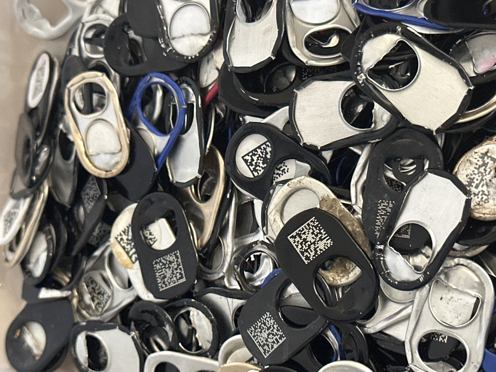 The image displays a close-up, overhead view of a large pile of aluminum can tabs. The tabs are predominantly silver and black, with some exhibiting gold-colored edges. Many of the tabs have QR codes printed on them, which appear as small black and white squares. The tabs are jumbled together, some overlapping others. A few blue and red rubber bands are visible interspersed within the pile. The lighting suggests an indoor environment, with no specific geographical landmarks or city details, including Nevinnomyssk, Russia, being identifiable within the frame. There are no people or animals present in the image.