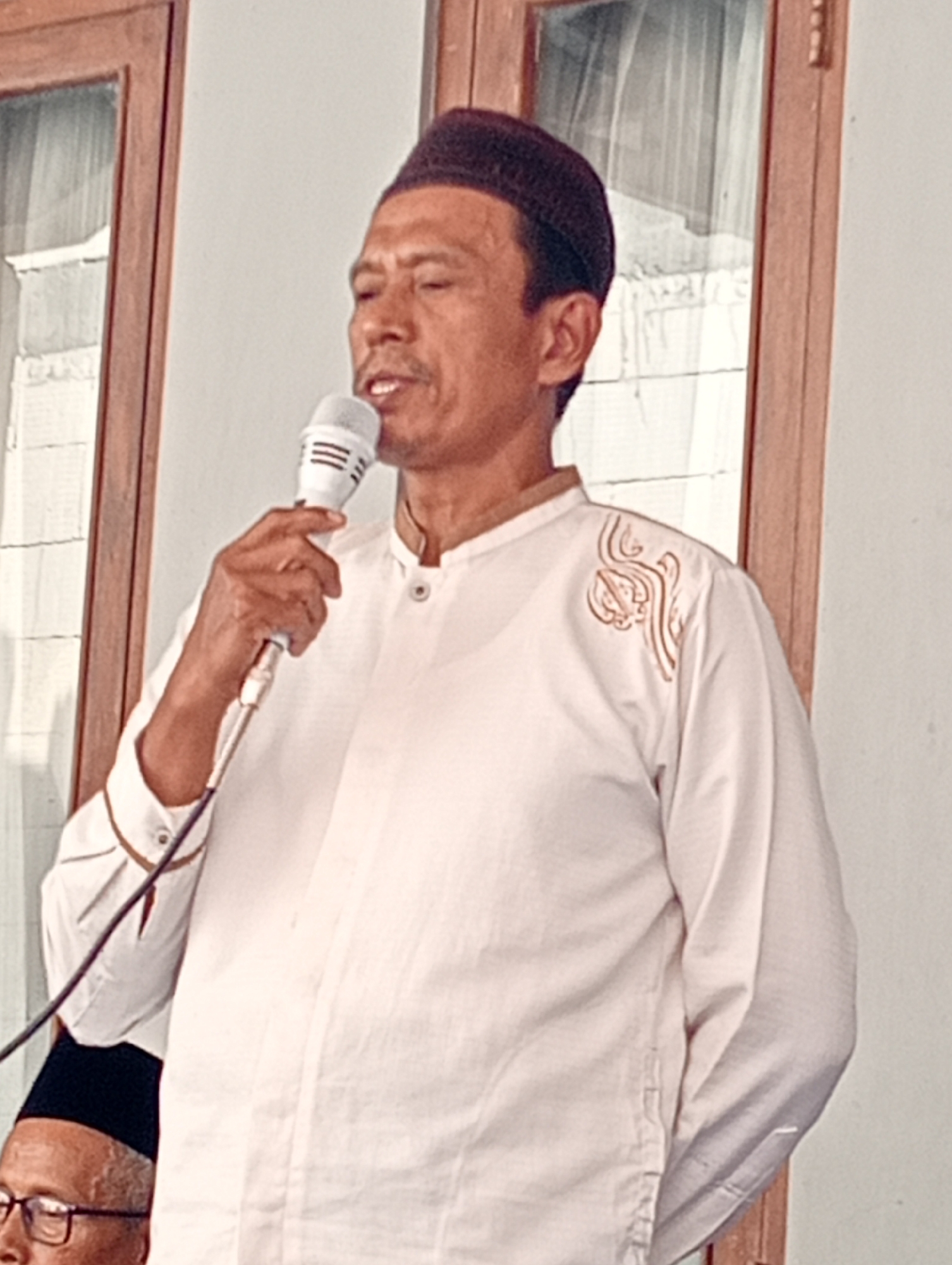 A man wearing a dark skullcap and a white long-sleeved shirt with gold embroidery on the left shoulder and cuffs is holding a white microphone and speaking into it. The man's eyes are closed and his mouth is open. He is standing in front of a wall and a wooden door with glass panels. The man's left hand is visible holding the microphone. Partially visible in the lower left corner is another person wearing glasses and a dark head covering. The image is taken in Boyolali, Indonesia, at the Alun-Alun Kidul, a public square, where a community event is taking place.