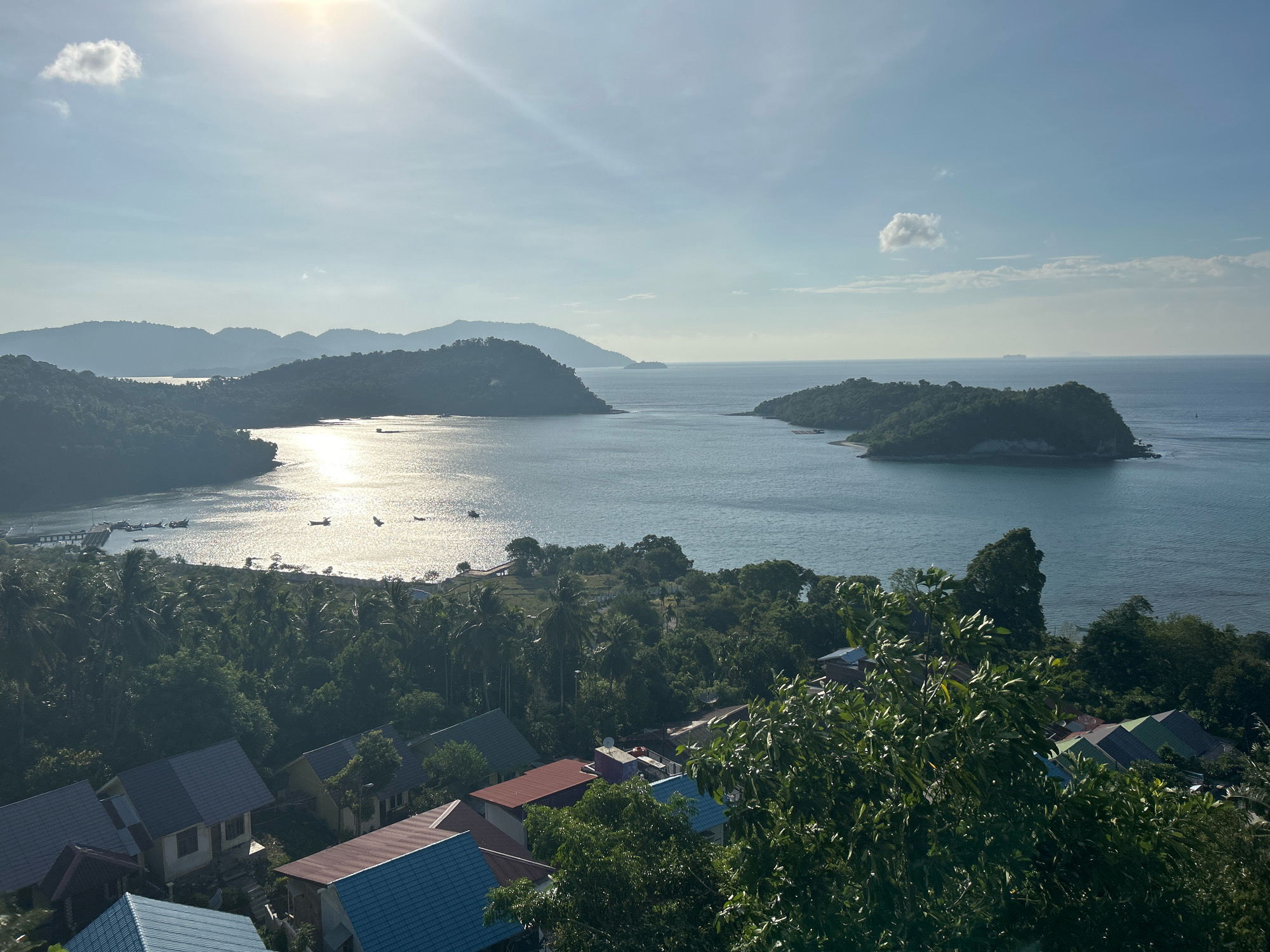 This is a scenic, elevated, wide-angle shot of a coastal area in Sabang, Indonesia. The sun is shining brightly, creating a shimmering effect on the ocean. In the foreground, there are several houses with colorful roofs, partially obscured by lush green trees. Beyond the houses, a dense jungle stretches towards the coastline. The sea is calm, with a few small boats visible in the water near a dock. In the mid-ground and background, several lush green islands and peninsulas rise from the ocean, with a hazy mountain range in the far distance. The sky is a clear blue with a few scattered white clouds, and the sun is a bright, hazy orb in the upper left quadrant of the image. The overall impression is of a tropical paradise bathed in sunlight.
