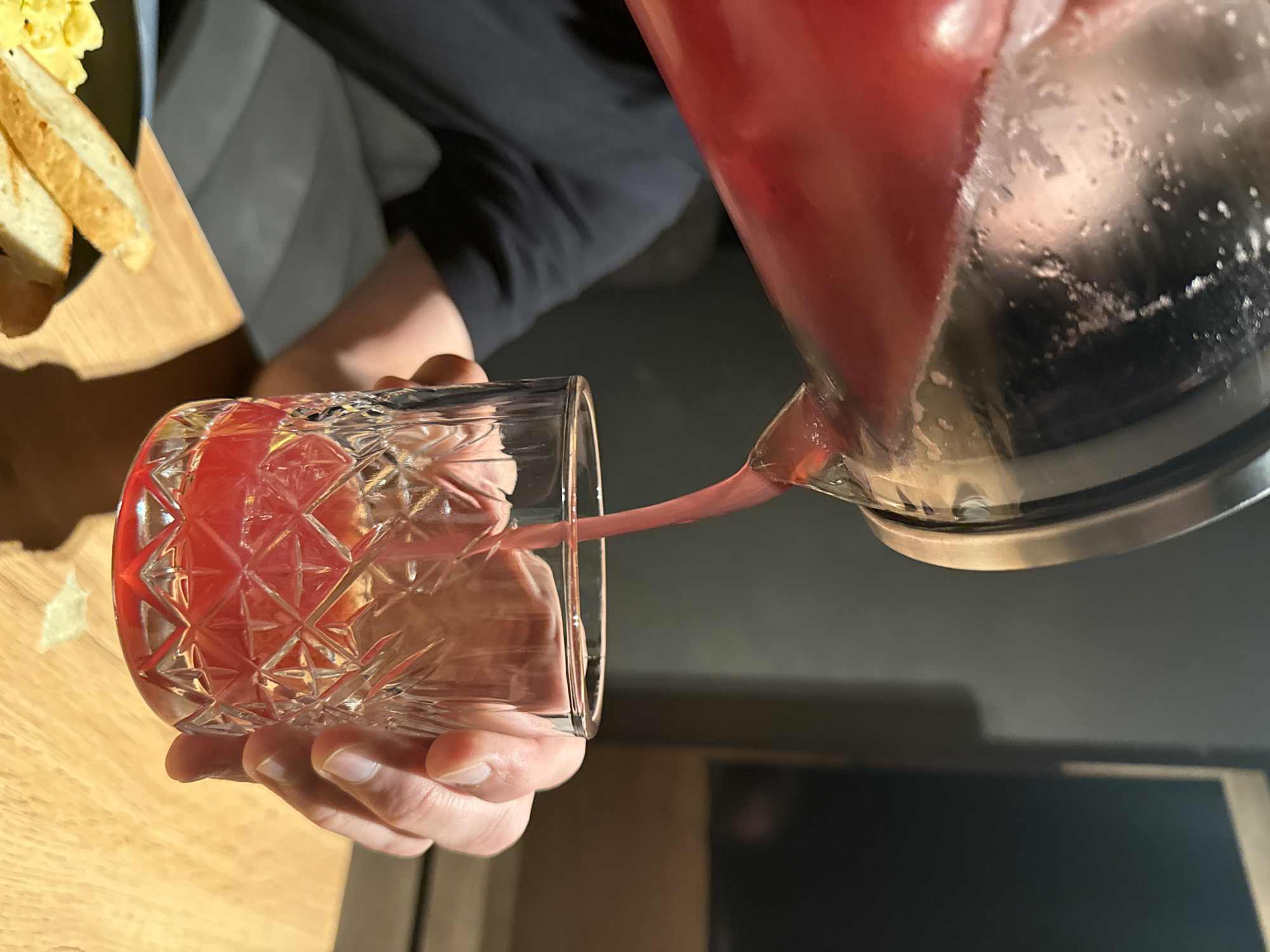 A person is pouring a reddish beverage from a clear pitcher into a cut-glass tumbler. The tumbler is partially filled with the liquid. A plate of food, possibly toast and scrambled eggs, is visible to the left. The background is a mix of light and dark surfaces. The location is not discernible from the image.